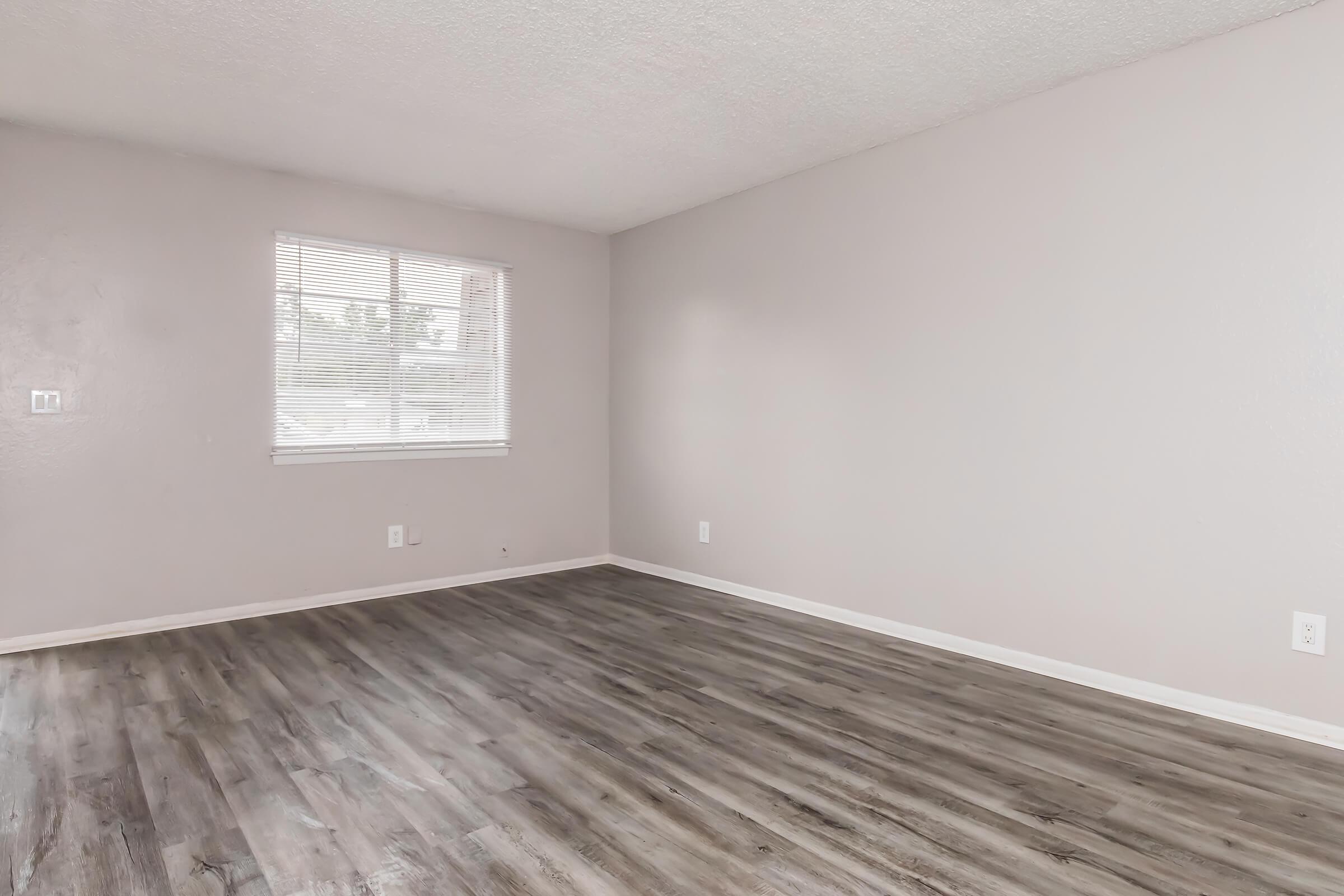 Empty room with light gray walls and a window with white blinds. The floor is covered with medium-tone laminate flooring. There are no furniture or decorations, creating a spacious and minimalistic atmosphere.