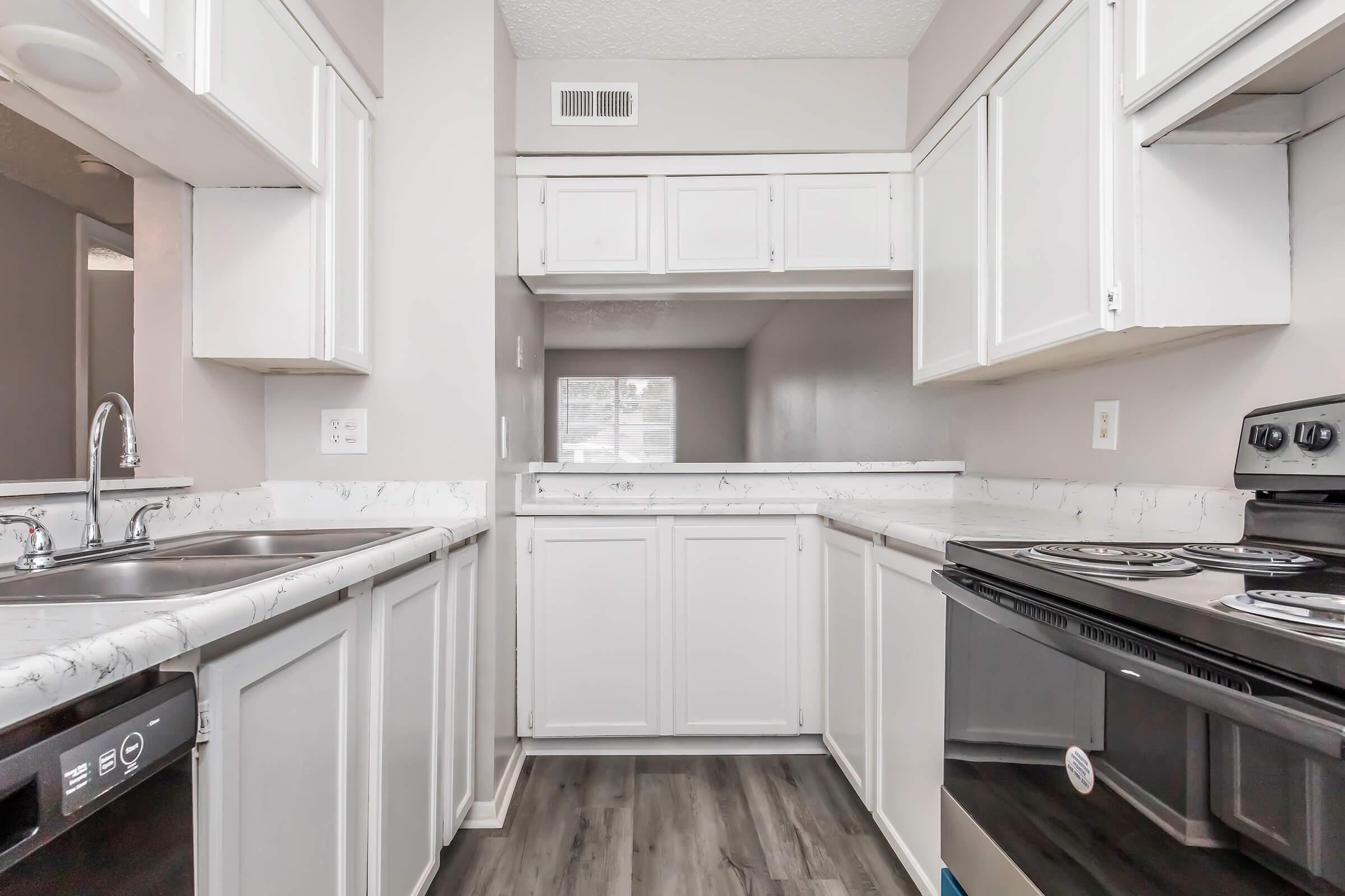 A modern kitchen featuring white cabinets, a double sink, and a black stove. The countertops have a marble design, and the flooring is dark wood. The kitchen has a spacious layout with an open design leading to a living area, and a window allows natural light to enter the space.