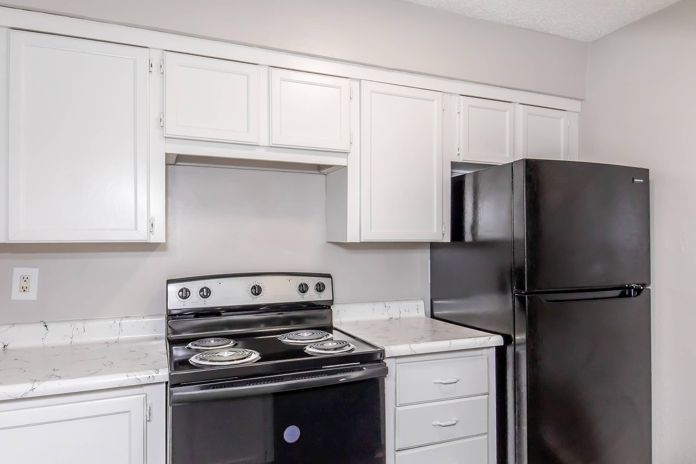 A modern kitchen featuring white cabinets, a black stove with four burners, a white countertop with a marble design, and a black refrigerator. The walls are a light gray, creating a clean and contemporary look.