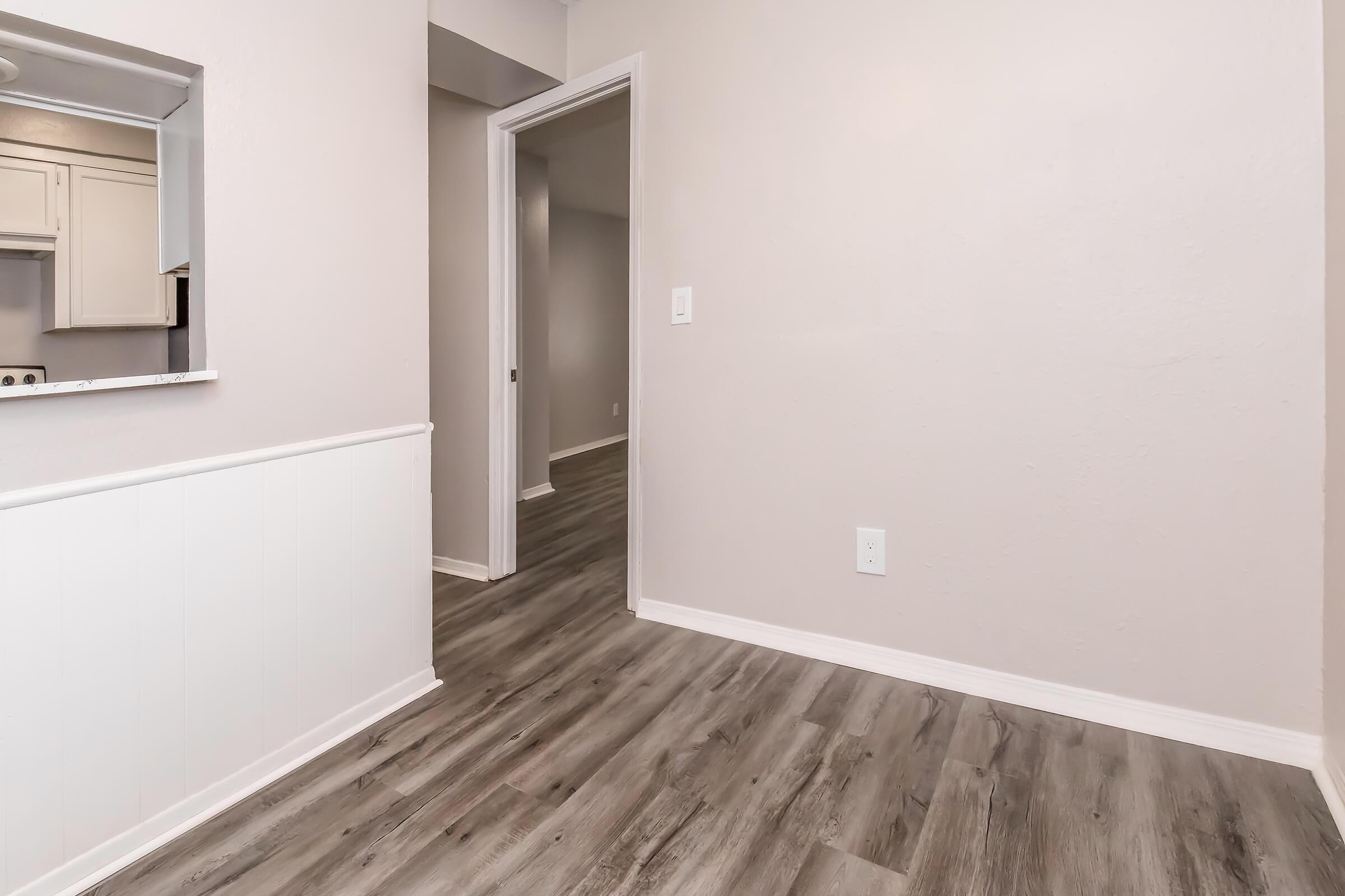A partially visible room featuring a light-colored wall, a wooden floor, and a doorway leading to another room. The left side shows a low wall with a small opening that appears to lead to a kitchen area. The overall atmosphere is bright and inviting, with neutral decor.