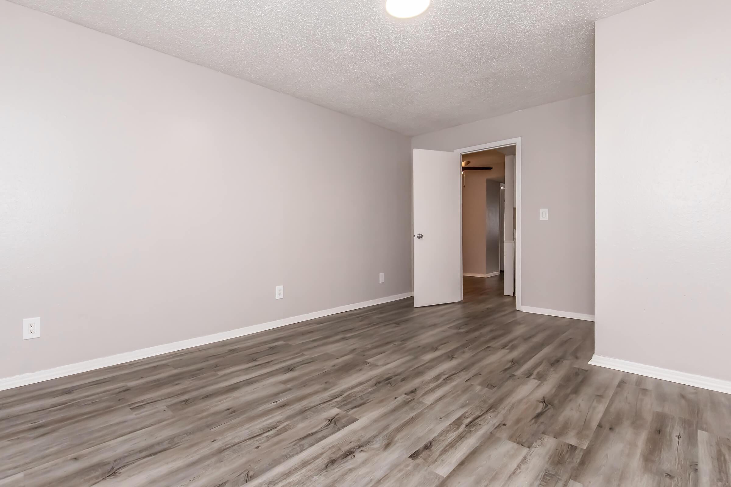 A spacious, empty room with light gray walls and wood-like vinyl flooring. A closed white door is visible on the right side, leading to another room. The ceiling has a soft white light fixture, providing a neutral and bright ambiance.