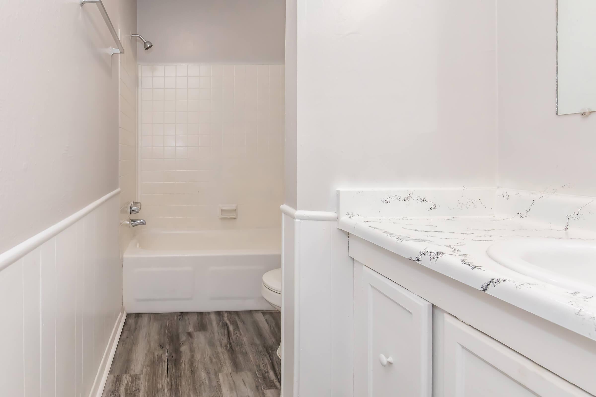 A clean and modern bathroom featuring a white bathtub with a shower, a toilet, and a countertop with a sink. The walls are painted light colors, and the floor has wooden planks. The area is well-lit and uncluttered, creating a fresh and inviting atmosphere.
