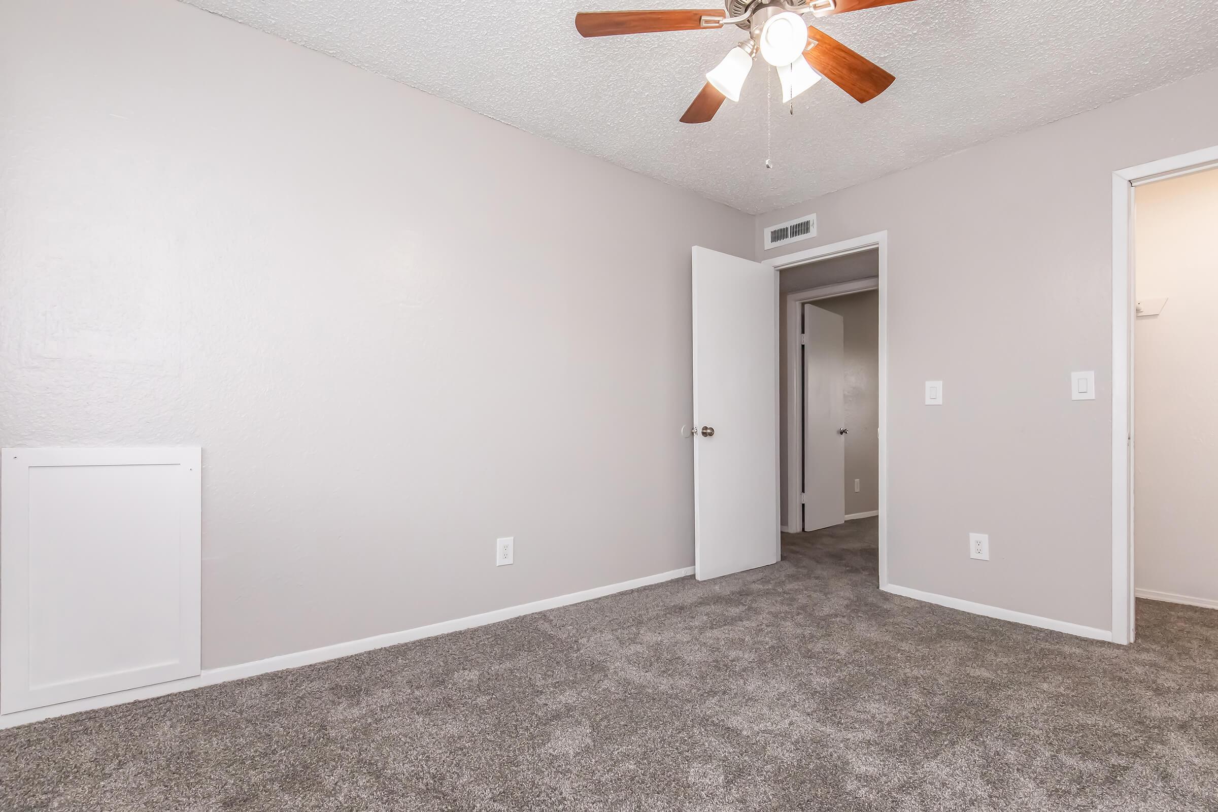 A spacious, neutral-colored bedroom with light gray carpet, a ceiling fan with wooden blades, and two doorways leading to other rooms. The walls are painted a soft beige, and there is a wall-mounted heating unit visible on the left. The room is well-lit, giving it a cozy and inviting atmosphere.
