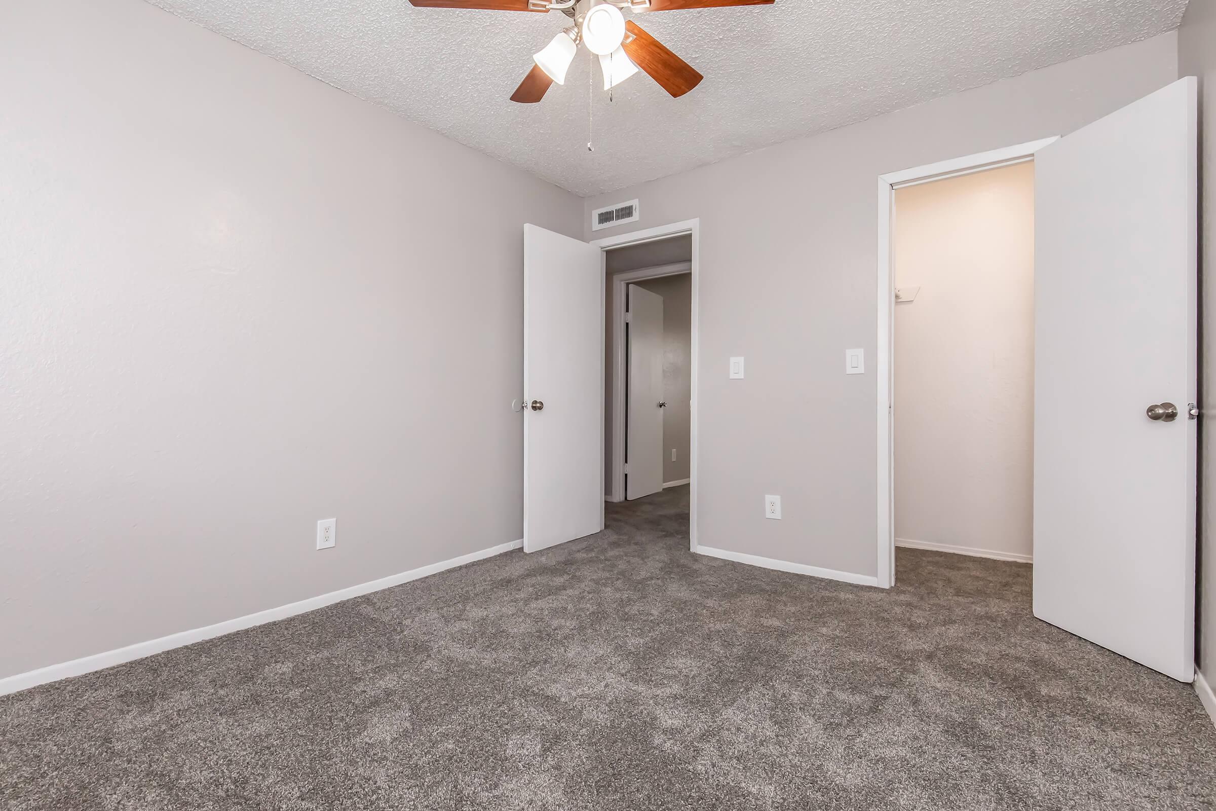 A small, empty room with light gray walls and plush gray carpet. There is a ceiling fan with wooden blades. Two white doors, one open and one closed, lead to adjoining spaces. The room is well-lit and has a neutral aesthetic, suitable for various decor styles.