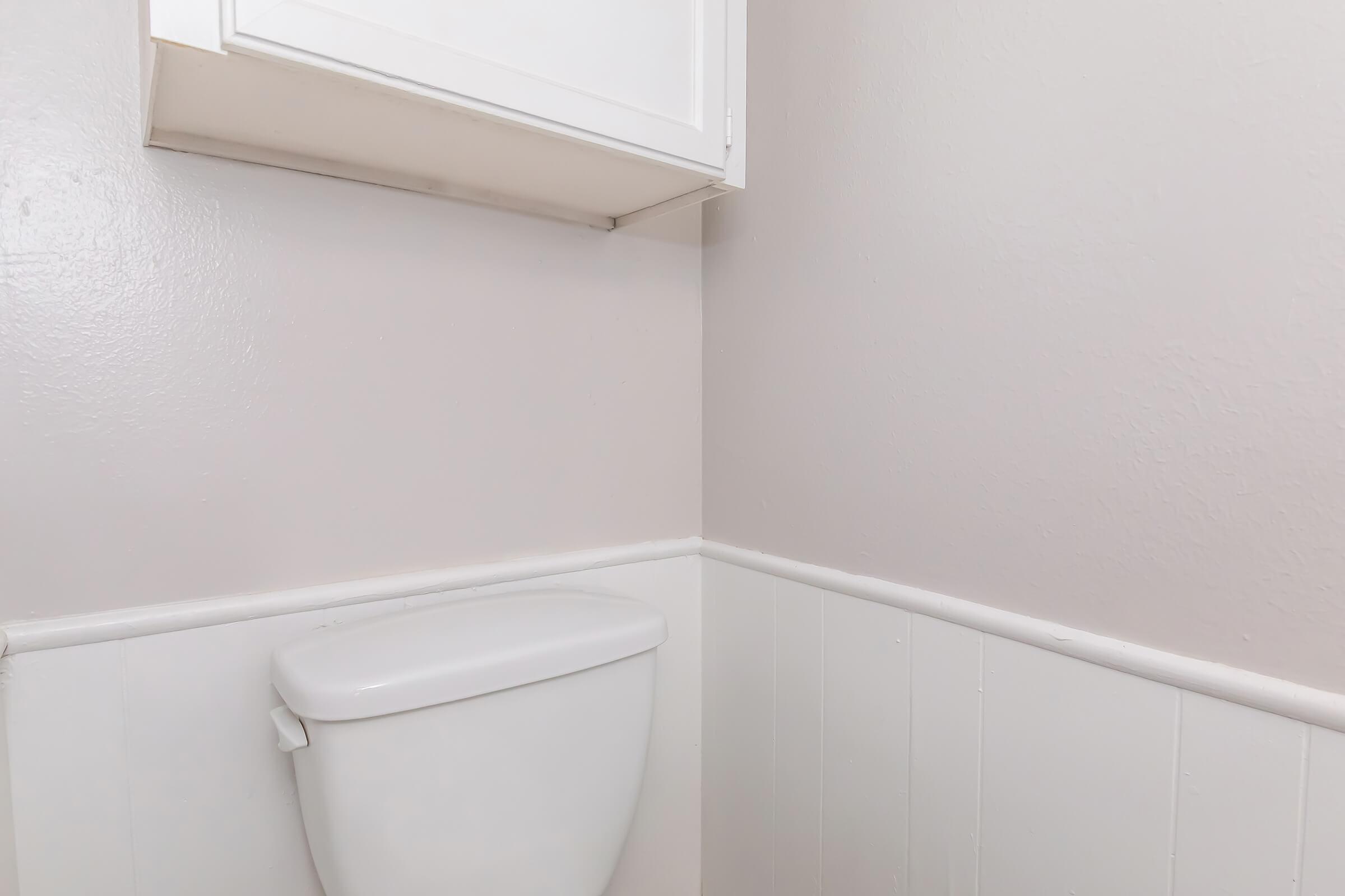 A corner of a bathroom featuring a white toilet tank and a light-colored wall. Above the toilet, there is a white cabinet mounted on the wall. The lower half of the wall has a wainscoting design in a matching light tone. The overall color scheme is neutral and minimalistic.