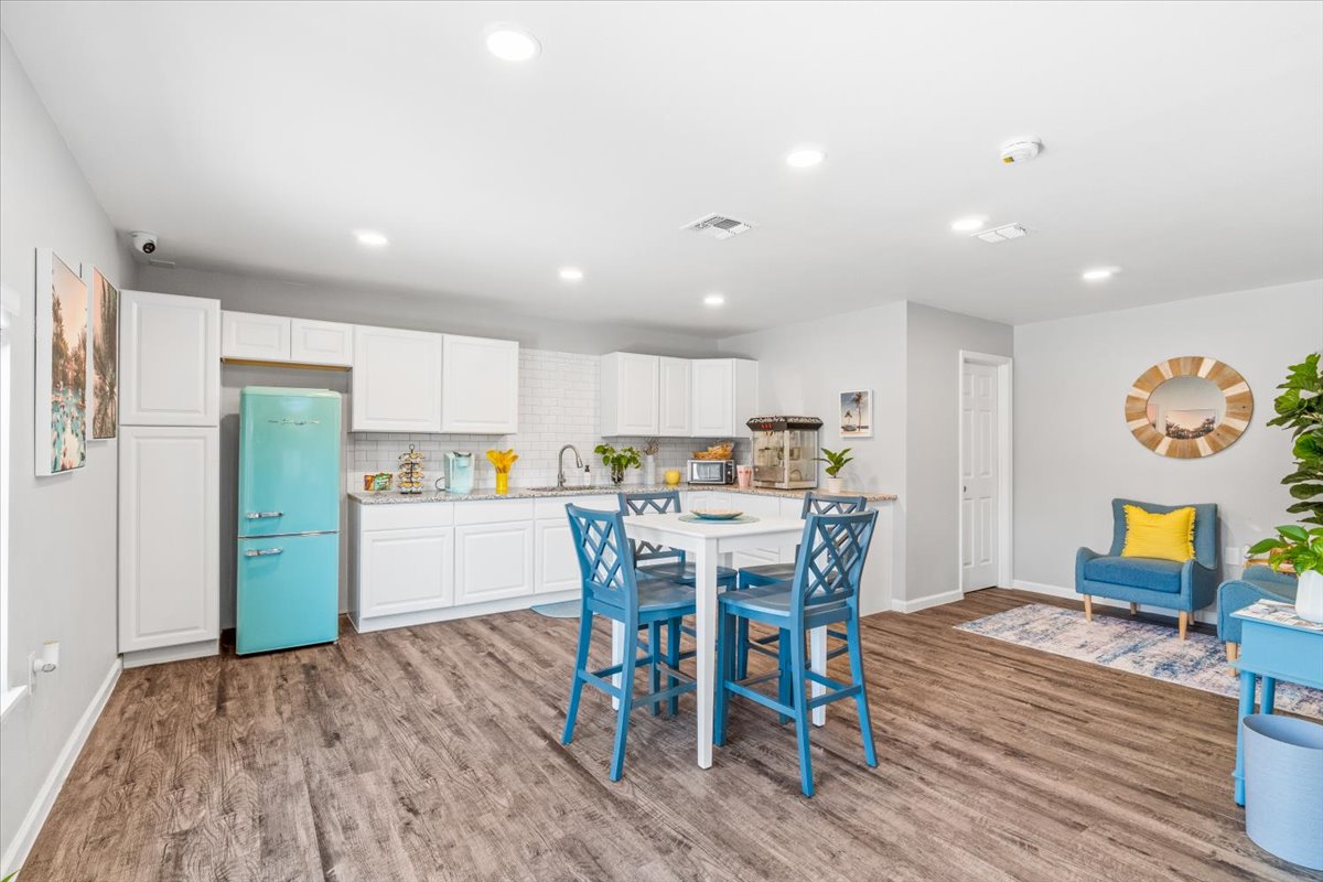 Modern kitchen and dining area featuring white cabinets, a retro turquoise refrigerator, a small dining table with blue chairs, and a cozy seating area with a blue armchair and yellow pillows. The space is well-lit with recessed lighting and has a wooden floor, creating a welcoming atmosphere.