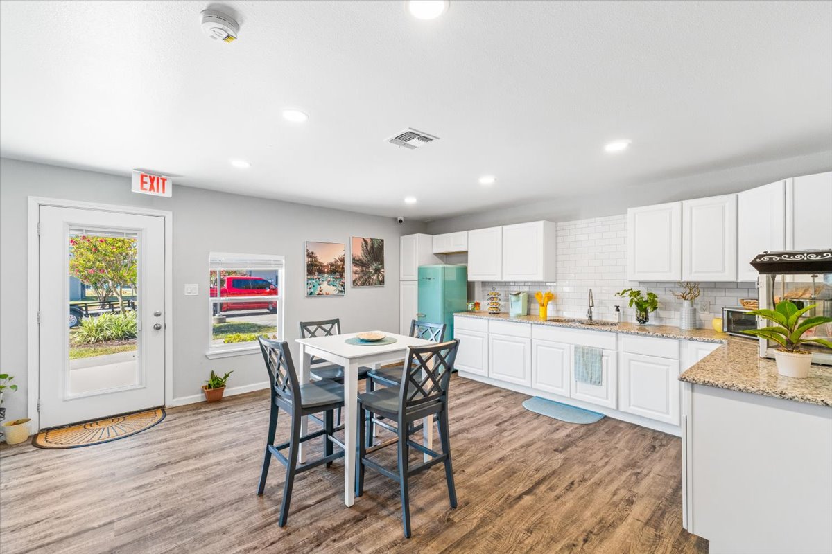 A bright and modern kitchen featuring white cabinetry, a granite countertop, and stainless steel appliances. A table with four chairs is in the center, surrounded by plants. Natural light comes through a door leading outside, with decorative artwork on the walls. The floor is made of brown laminate planks.