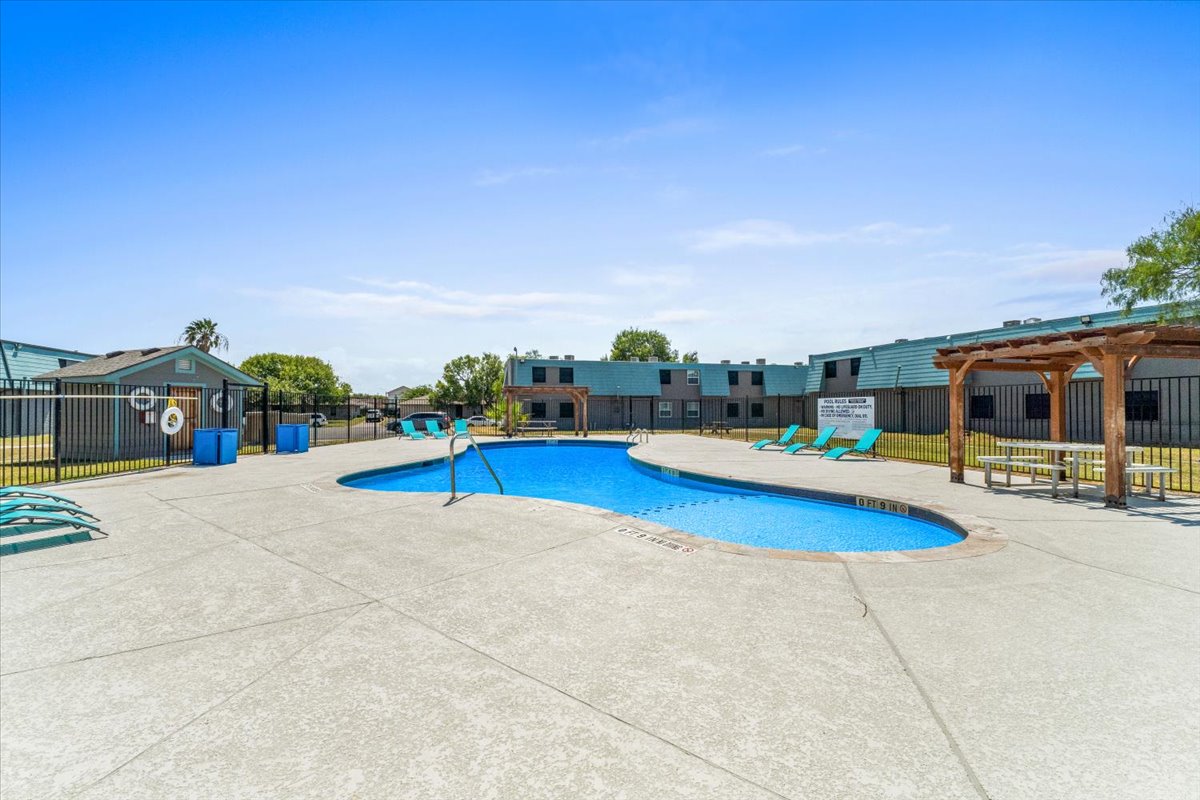 A clear blue swimming pool surrounded by a spacious patio area with lounge chairs. In the background, there are residential buildings and greenery. A pergola is visible near the pool, and the area is enclosed by a fence. The sky is bright and sunny, indicating a warm day.