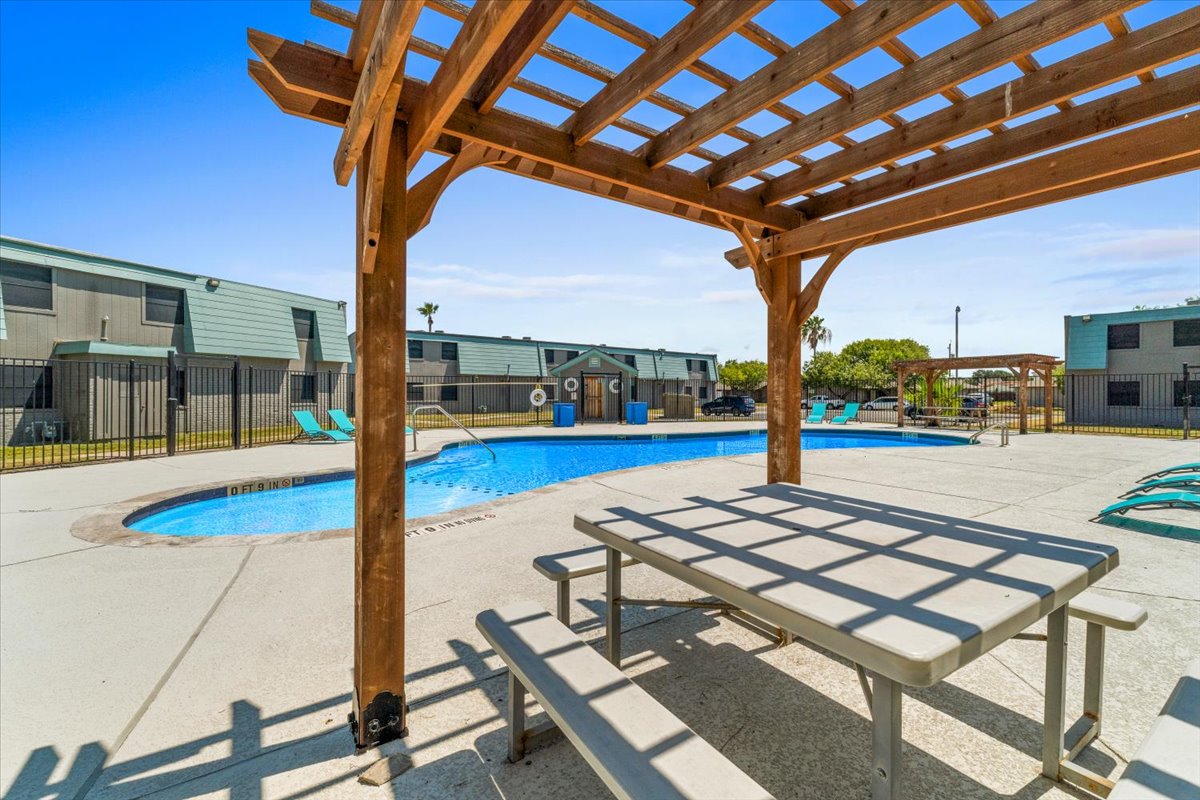 A wooden pergola provides shade over a picnic table near a swimming pool. The pool is surrounded by lounge chairs, and there are buildings in the background under a clear blue sky. The area is well-maintained, creating a relaxed outdoor atmosphere.