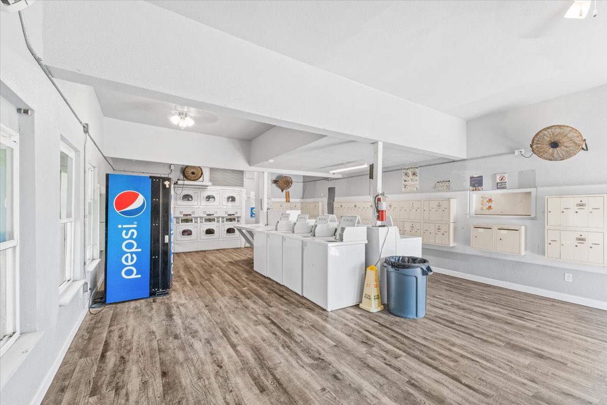 A bright, spacious laundry room featuring several washing machines and dryers, surrounded by light-colored walls and wooden flooring. On one side, there’s a Pepsi vending machine, while the other side displays various mailboxes. The area is clean and well-organized, with good natural light illuminating the space.