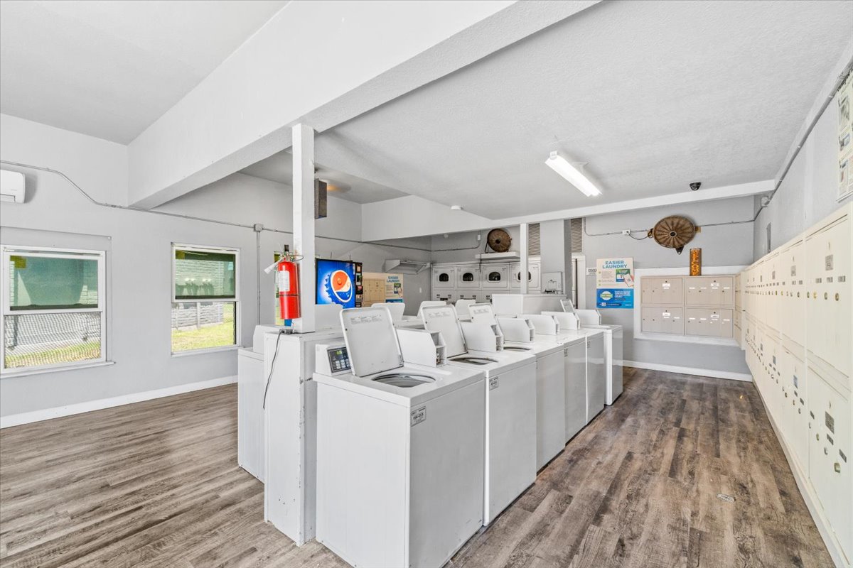 A clean and bright laundry room featuring several washing machines and dryers arranged in a row. There are lockers on the right side, a fire extinguisher mounted on the wall, and a television displaying a logo. The flooring is wood-like, and there are windows allowing natural light into the space.