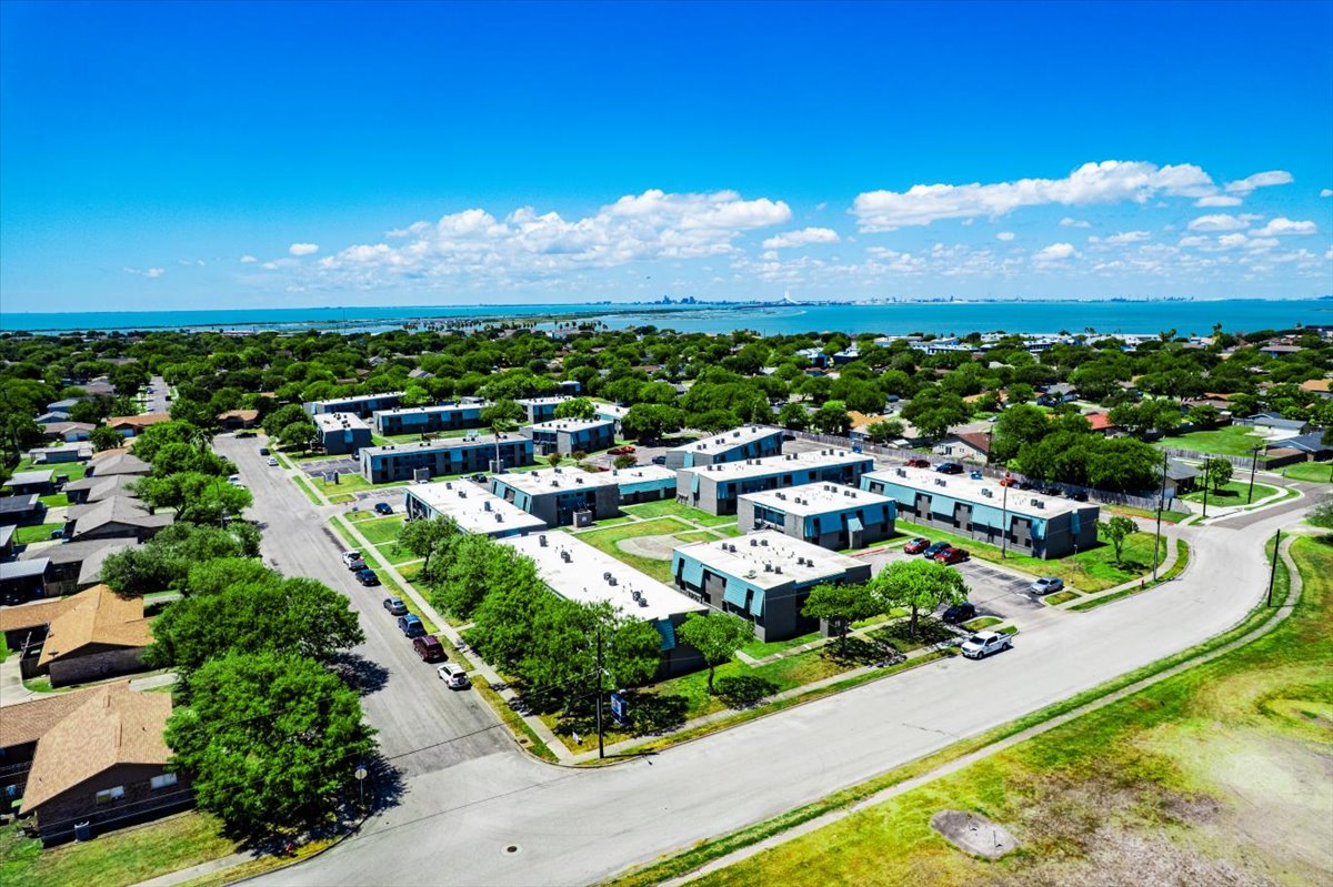 Aerial view of a residential neighborhood featuring low-rise buildings surrounded by greenery, with a clear blue sky and distant views of a city skyline along the coastline. The scene includes roads, parking areas, and a mix of homes with landscaped lawns.