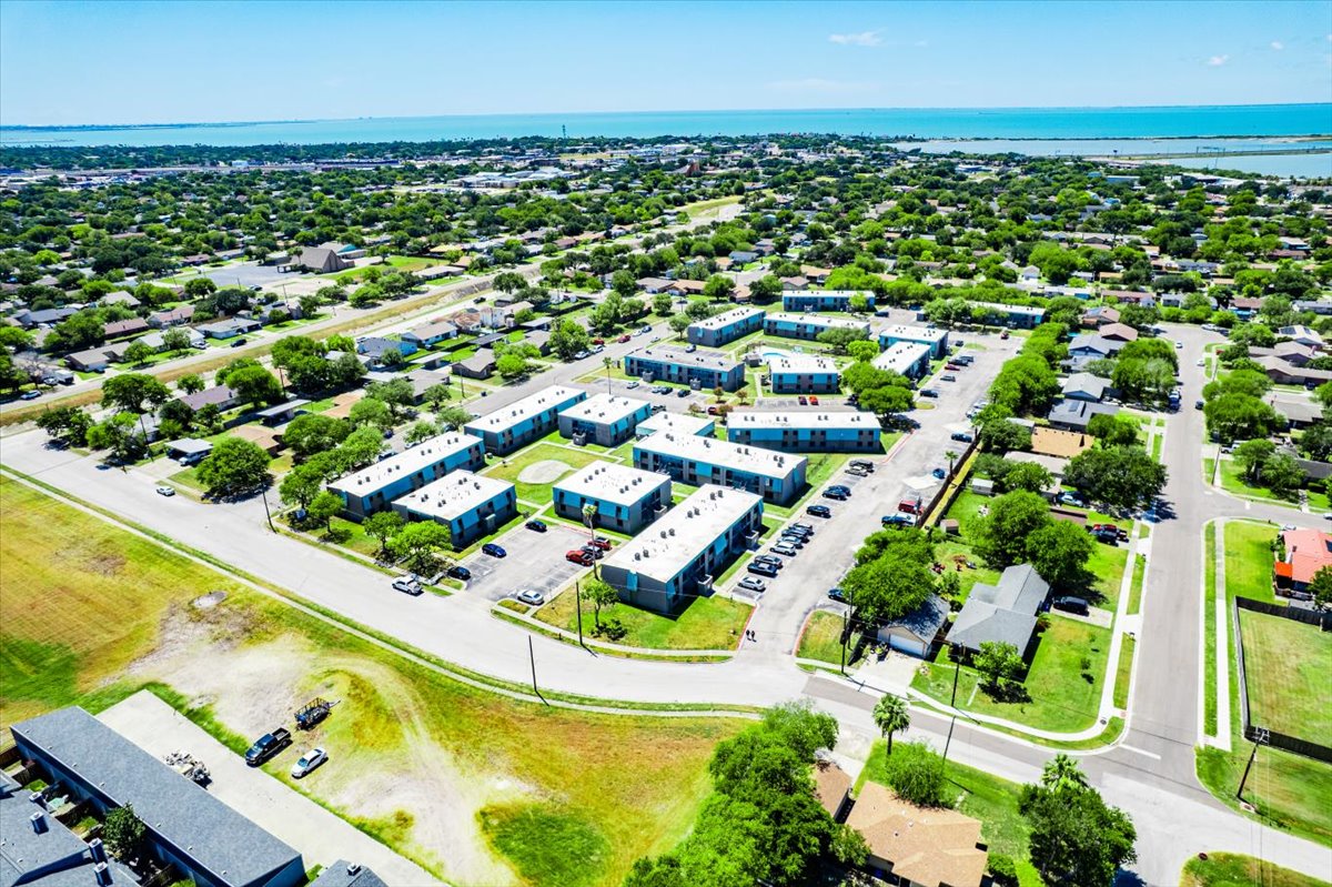 Aerial view of a residential area featuring clusters of low-rise apartment buildings surrounded by green spaces and roads. In the background, a body of water is visible, with more houses and greenery in the vicinity, indicating a suburban environment.