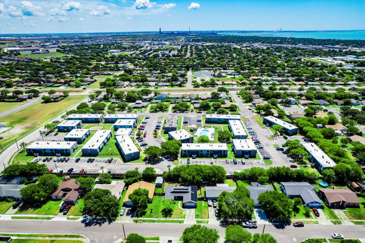 Aerial view of a suburban neighborhood featuring several residential buildings arranged in clusters. The area is lush with greenery, including trees and lawns, with a mix of single-family homes and apartment complexes. In the background, the horizon shows distant industrial buildings and a body of water.
