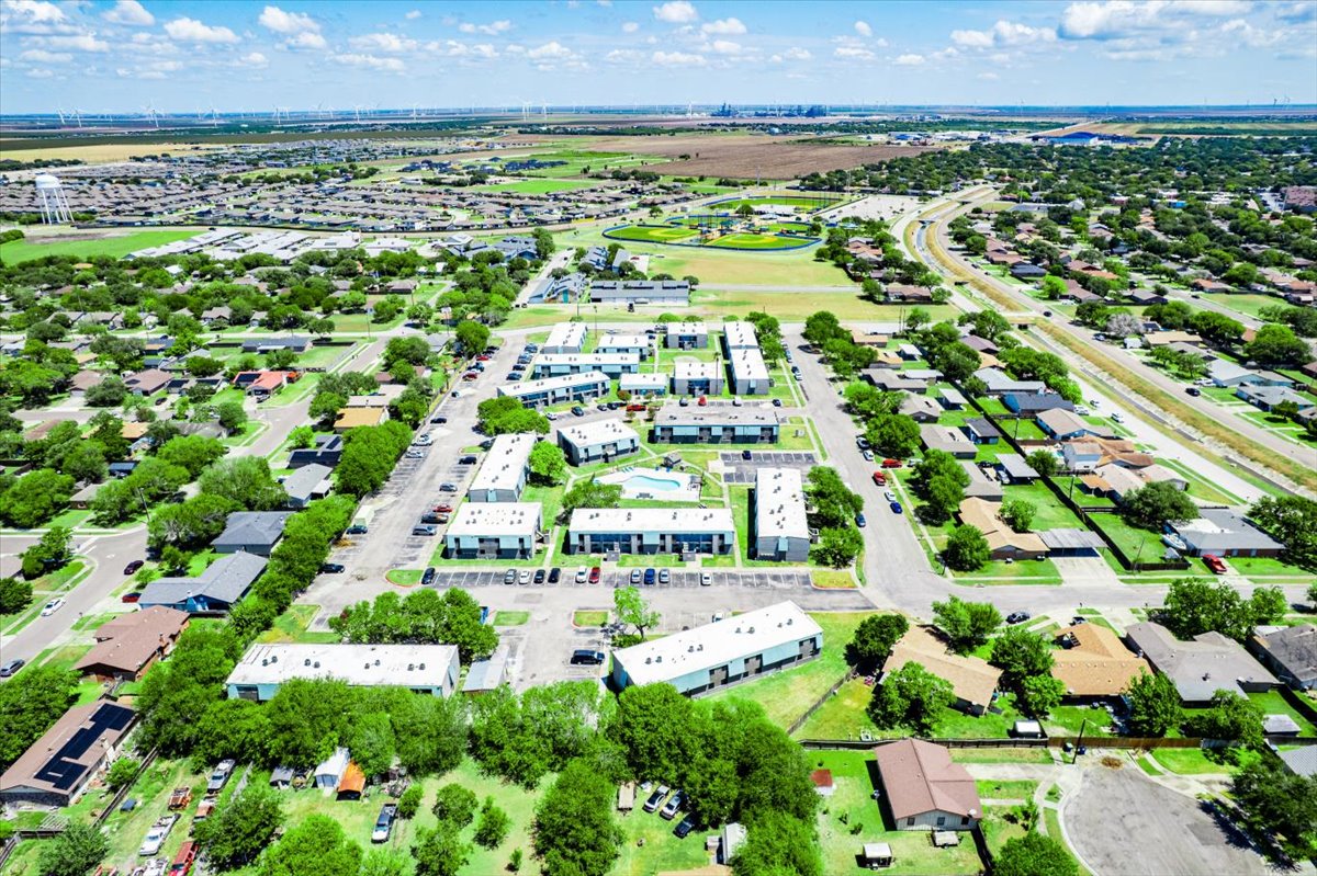 Aerial view of a suburban neighborhood with a mix of residential buildings and green spaces. The landscape features clusters of houses, roads, and patches of trees. In the distance, wind turbines and open fields are visible, alongside a highway. The sky is partly cloudy, creating a vibrant and expansive scene.