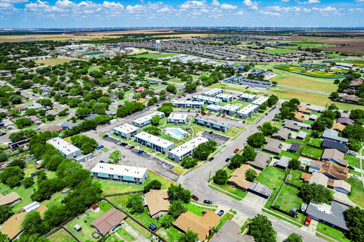 Aerial view of a suburban neighborhood featuring clusters of residential buildings, a swimming pool, and well-maintained lawns. In the background, there are fields and wind turbines, with a clear blue sky. Various cars are parked along the streets that wind through the area.