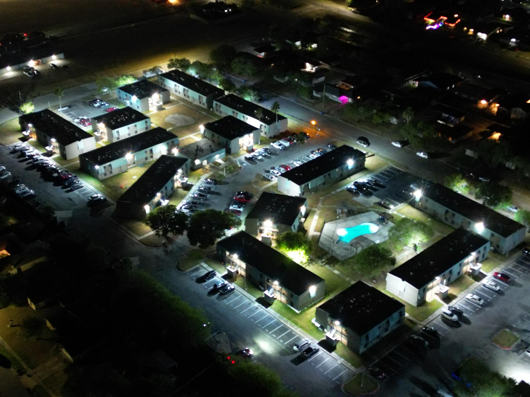 Aerial view of a residential complex at night, featuring multiple two-story buildings arranged in a U-shape. There are parked cars in designated spaces, and a swimming pool is illuminated in the center of the complex. Surrounding areas are dimly lit with some streetlights and nearby buildings visible.