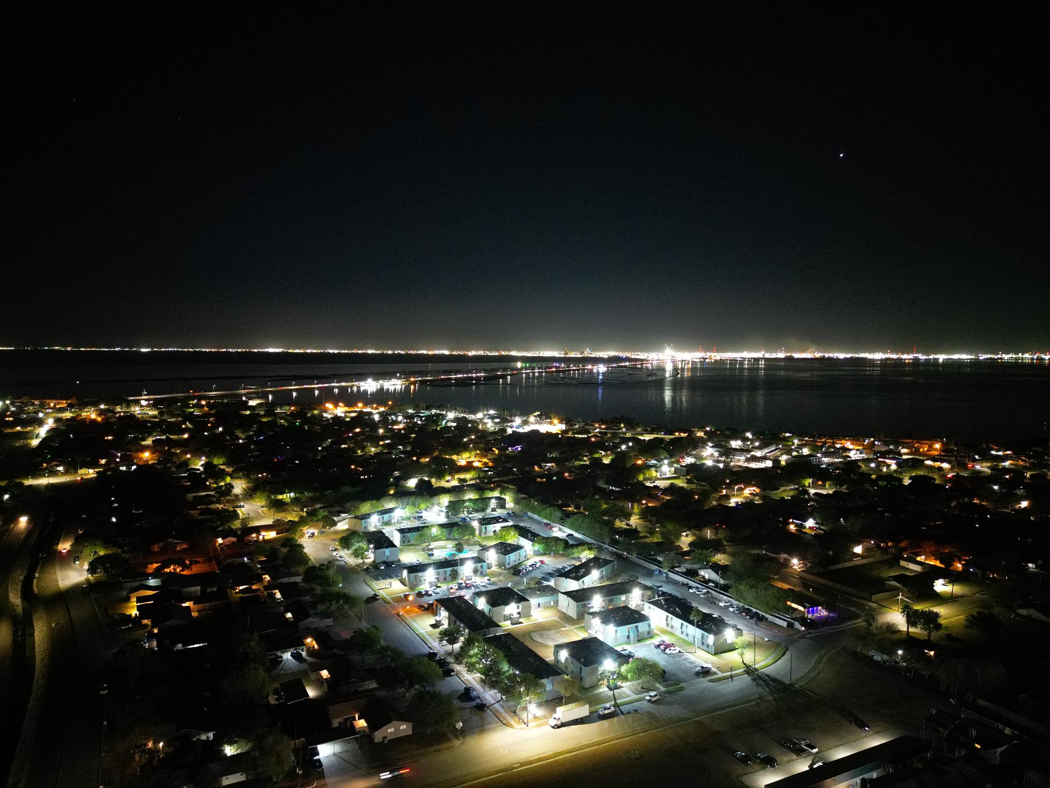 Aerial view of a coastal city at night, showcasing brightly lit neighborhoods and streets. The shoreline glimmers with lights reflecting on the water, while a distant bridge and cityscape can be seen across the bay. The scene captures a peaceful night atmosphere with scattered illuminated buildings.