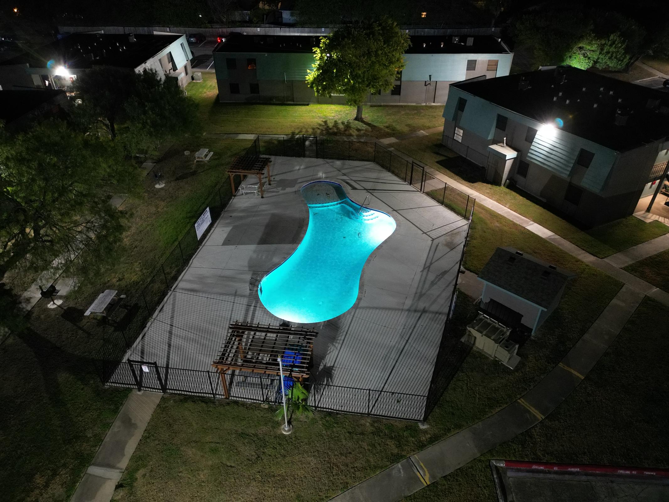 Aerial view of a swimming pool in a residential area at night. The pool has an irregular shape and is illuminated by soft blue lights. Surrounding the pool are fences and landscaped greenery, with nearby buildings visible in the background. Pathways and seating areas are also present around the pool area.