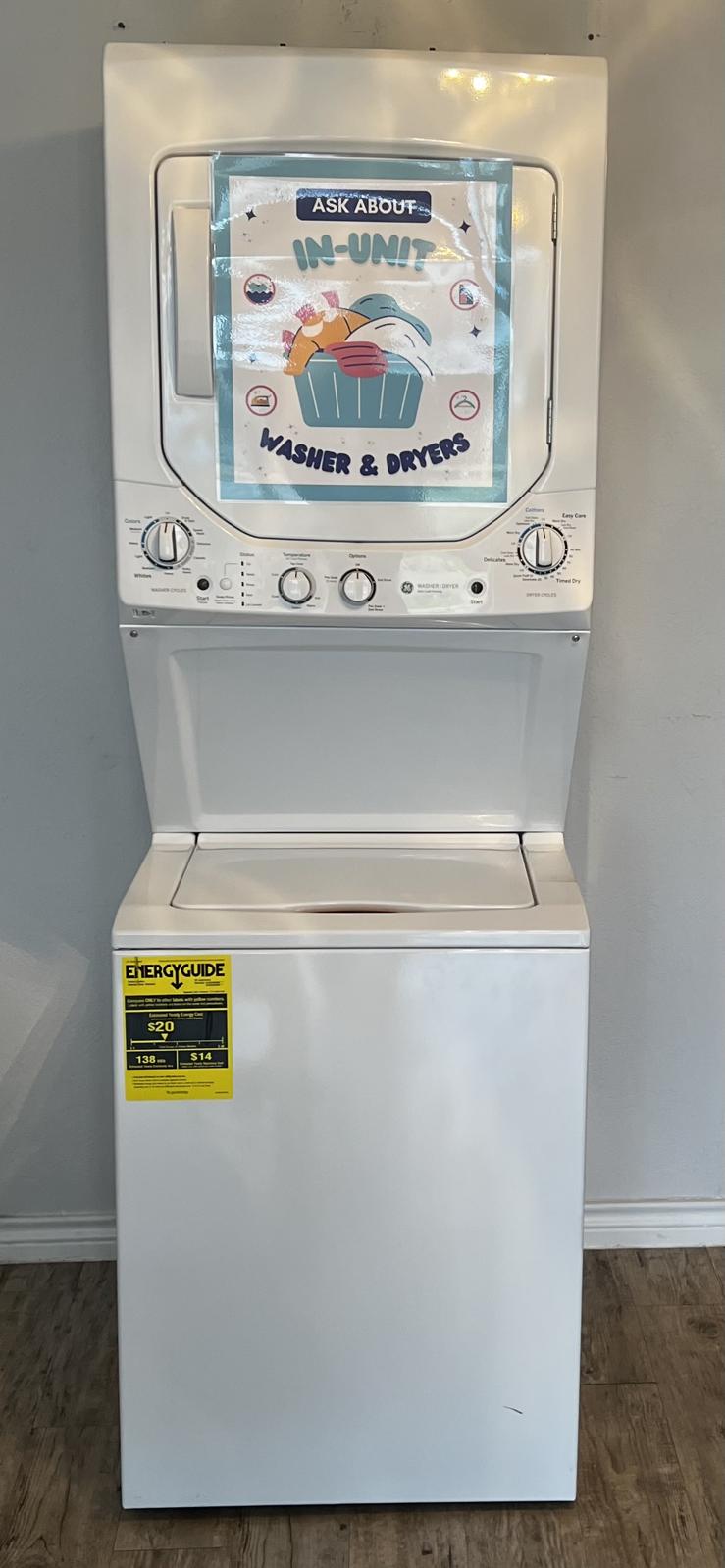 A stacked washer and dryer unit in a neutral-colored room. The washer and dryer are white, with a sign above them that reads "ASK ABOUT IN-UNIT WASHER & DRYER." A bright yellow energy guide sticker is affixed to the front of the unit. The floor is wooden, adding a warm touch to the space.