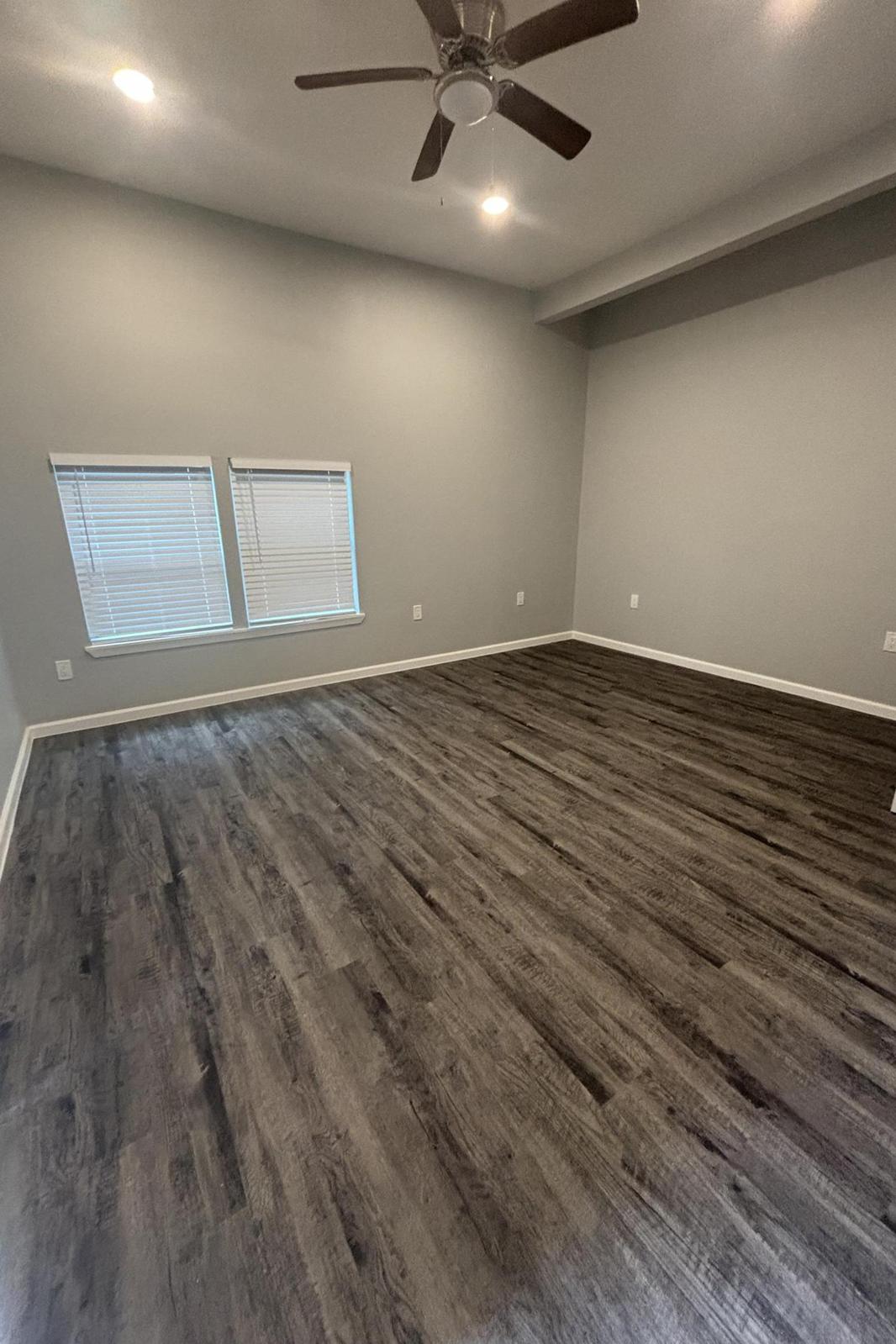 Interior of a spacious room with gray walls and dark wood-like flooring. There are two windows with blinds allowing natural light to enter. The ceiling features a ceiling fan, and the room is empty, giving it an open and airy feel.