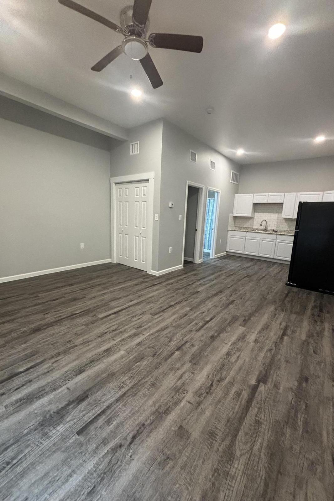 A spacious, modern living area featuring light gray walls, a ceiling fan, and laminate flooring. There's a doorway leading to a kitchen with white cabinets and a black refrigerator in the corner. The room is bright with natural light coming from windows in the distance.