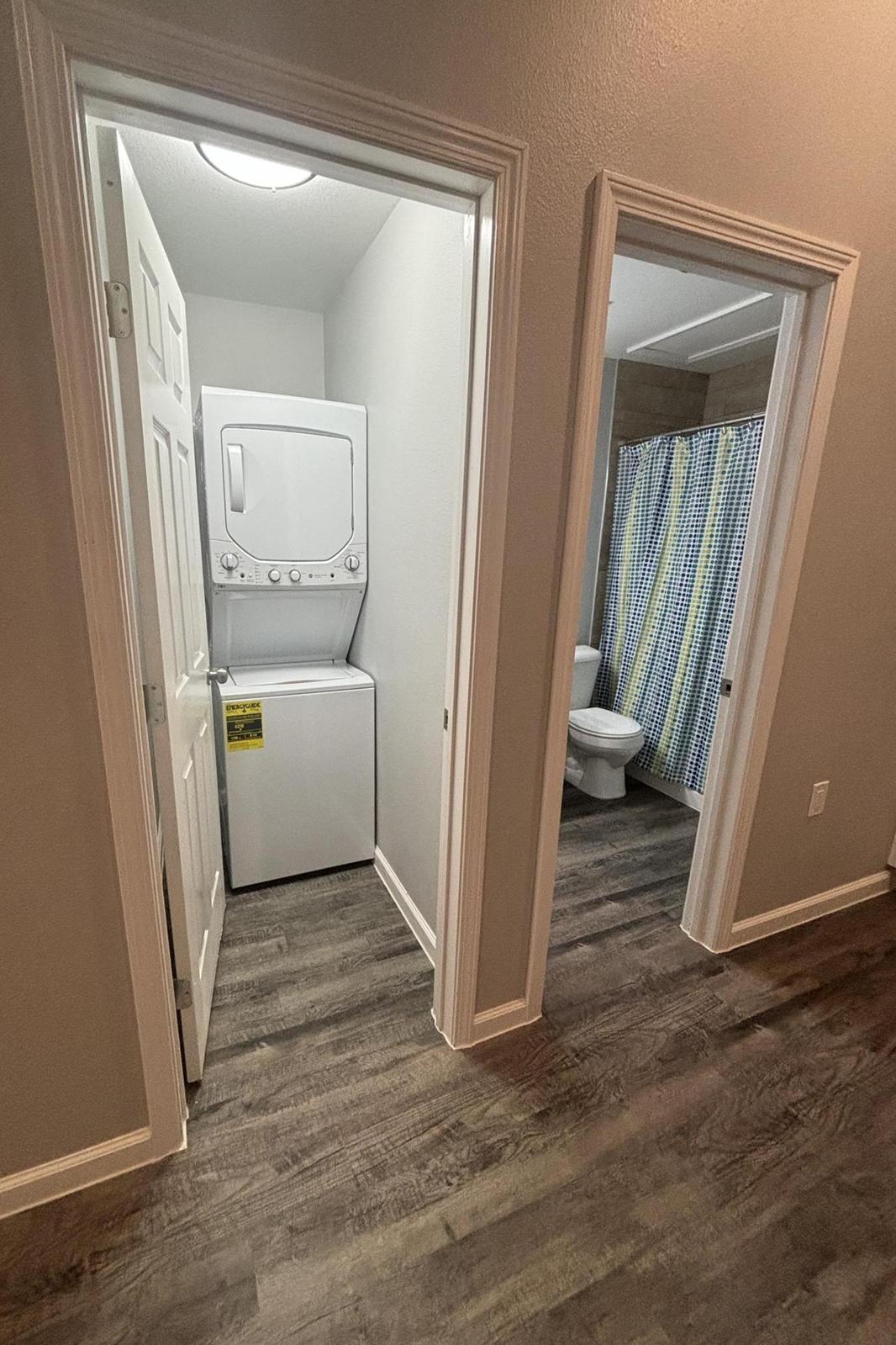 A view of a laundry area with a stacked washer and dryer on the left, and a bathroom with a shower curtain in blue and yellow stripes on the right. The walls are a light gray, and the flooring is a dark wood finish. Two doors lead into the laundry and bathroom spaces, respectively.
