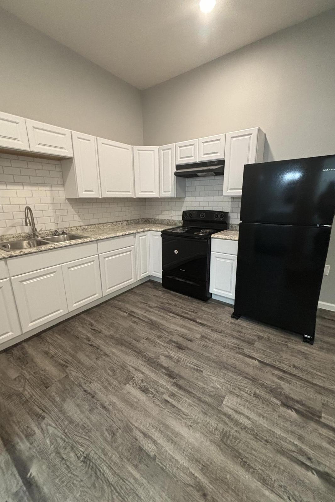 A modern kitchen featuring white cabinetry, a granite countertop, a stainless steel sink, a black stove, and a black refrigerator. The flooring is a light wood tone, and the walls are painted in a neutral color. The kitchen is well-lit with overhead lighting.