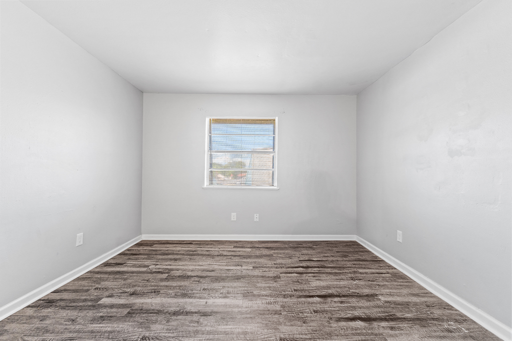 Empty room with light gray walls and a wood-like floor. A single window allows natural light to enter, featuring blinds. The space is uncluttered, creating a minimalist and airy atmosphere.