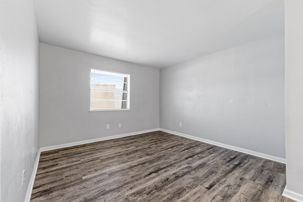 A vacant room with light gray walls and a window allowing natural light. The floor features a wood-like laminate, and the space appears clean and unfurnished, offering a blank canvas for decoration or use.