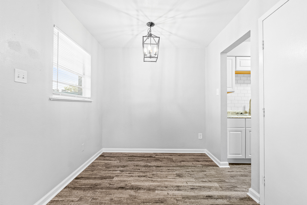 A bright, empty room with white walls and a wooden floor. A modern pendant light hangs from the ceiling, casting patterns on the wall. There is a window on the left, allowing natural light in, and an open doorway leading to a kitchen area, suggesting a spacious and inviting living environment.
