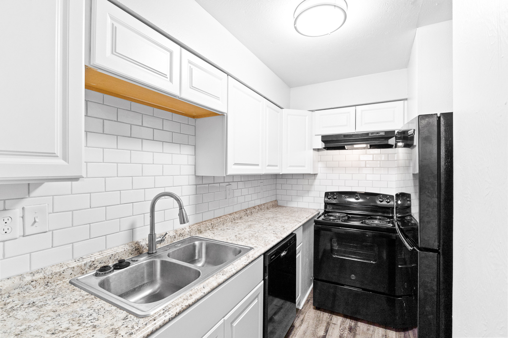 Modern kitchen featuring white cabinetry, a granite countertop, and a double sink. The kitchen is equipped with a black refrigerator, stove, and microwave, complemented by a white subway tile backsplash. Soft overhead lighting illuminates the space, creating a clean and bright atmosphere.
