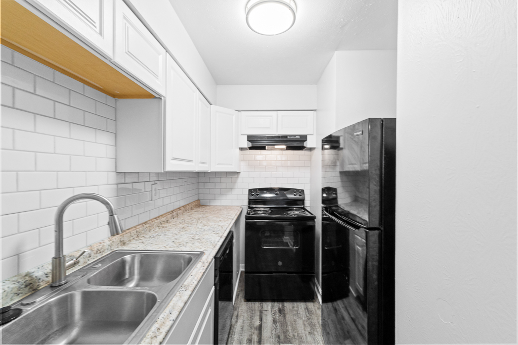 A modern kitchen featuring white cabinets, a granite countertop, and stainless steel appliances. The kitchen includes a double sink, a black stove, and a refrigerator, with white subway tile backsplash. A ceiling light provides bright illumination in a compact space.