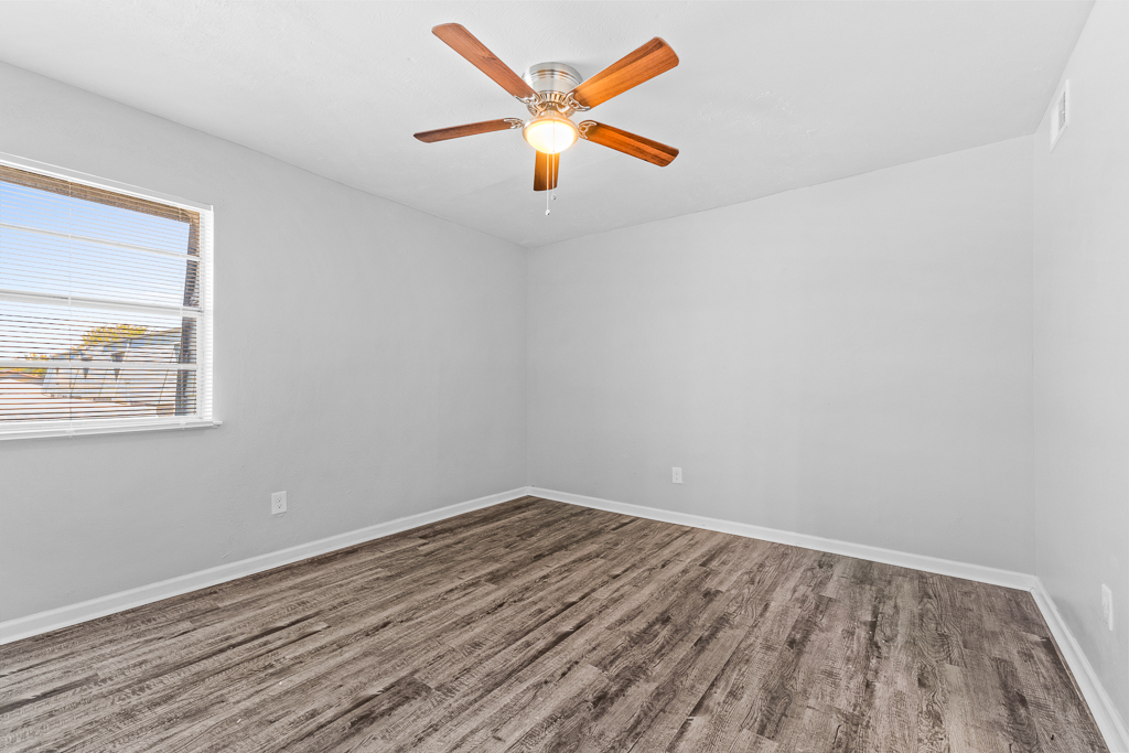 Empty room with light gray walls and a ceiling fan featuring wooden blades. Natural light streams in through a window with blinds. The flooring is a light-colored wood laminate, and there are no furnishings or decorations in the space, creating a clean and minimalist look.