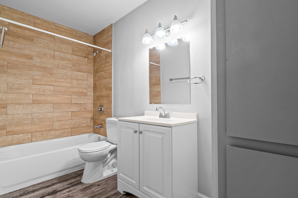 A modern bathroom featuring a bathtub, a white toilet, and a vanity with a sink. The walls are adorned with light wooden panels and the area is illuminated by a stylish light fixture above the mirror. The flooring is a wood-like finish, creating a warm and inviting atmosphere.