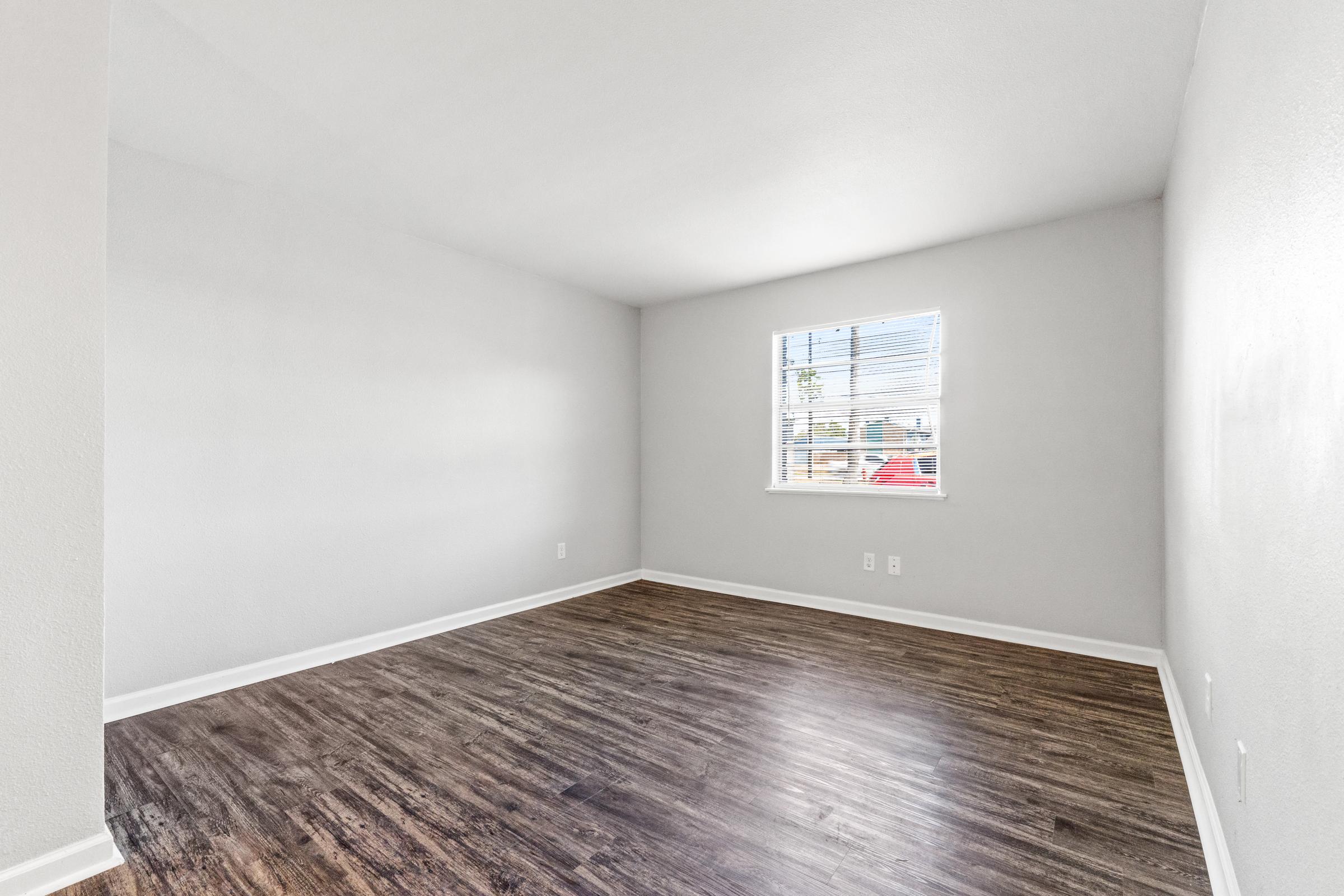 A spacious, empty room with light gray walls and wooden flooring. A window on one wall allows natural light to brighten the space, and there are no furnishings or decorations, creating a clean and minimalist appearance.