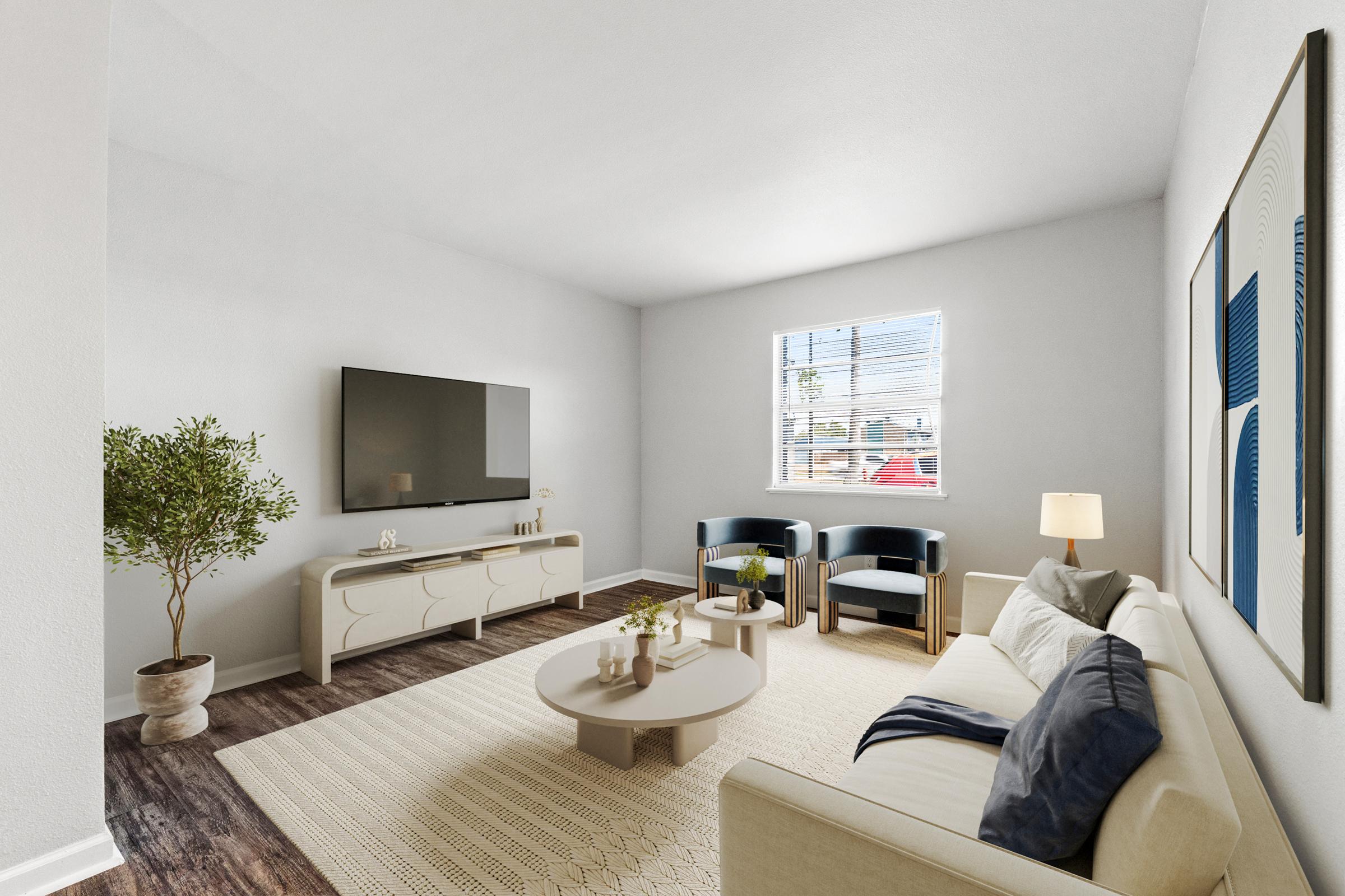 A modern living room featuring a light-colored couch with a navy throw pillow, a round coffee table, and two dark blue armchairs. A flat-screen TV is mounted on the wall opposite the seating area. A potted plant adds greenery, while bright natural light filters through a window, highlighting the neutral decor.