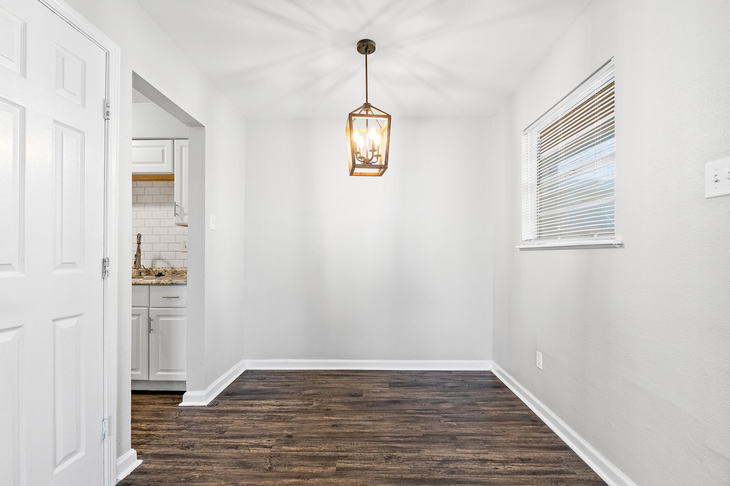 Interior view of a well-lit room featuring a hanging lantern light fixture, light gray walls, and dark wood flooring. A doorway leads to a kitchen area with tile backsplash visible. A window allows natural light to enter, creating a bright and inviting atmosphere.