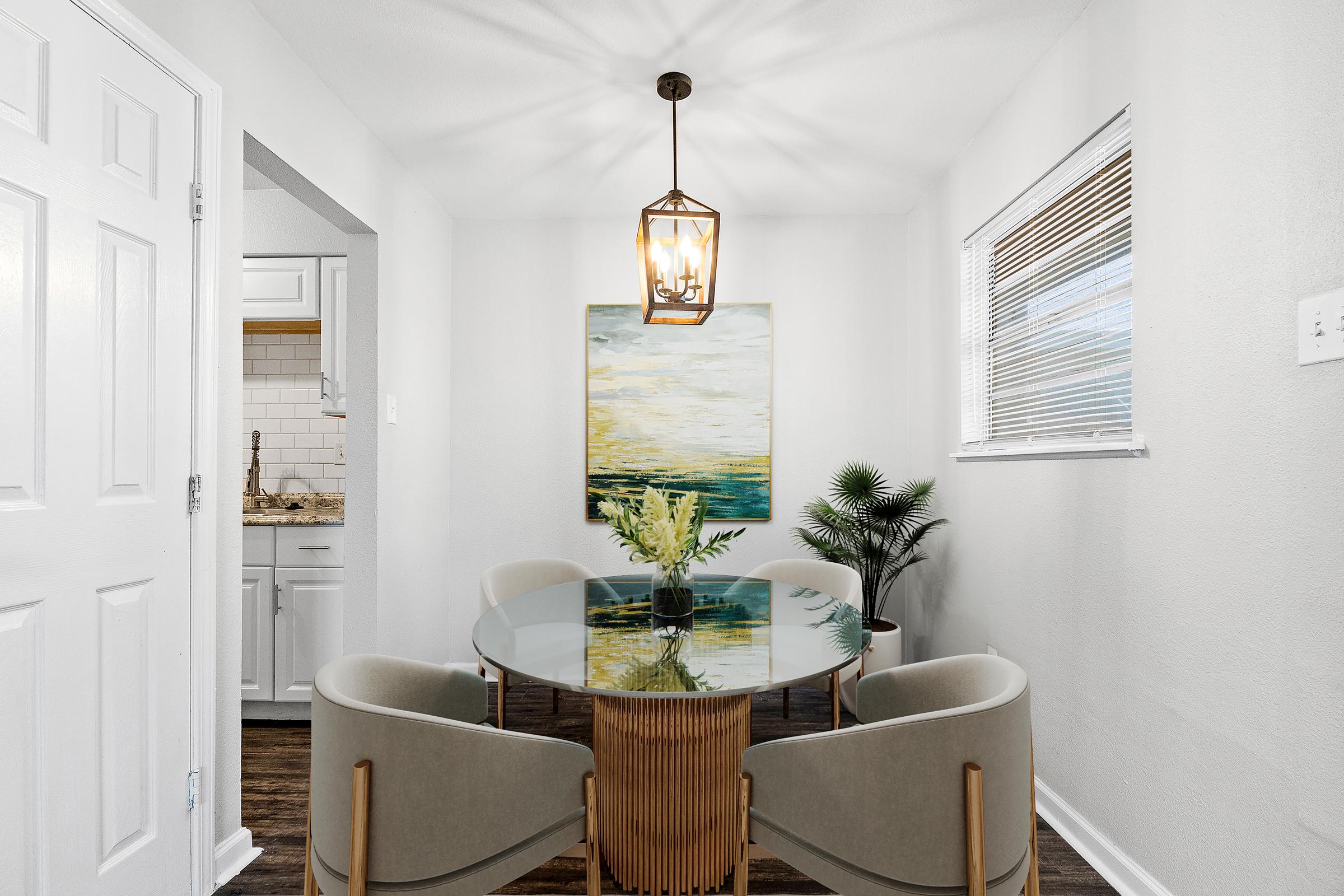 A modern dining area featuring a round glass table surrounded by four upholstered chairs. A pendant light hangs above the table, and a large abstract painting is mounted on the wall. A potted plant adds a touch of greenery, while the room has white walls and polished wooden flooring.