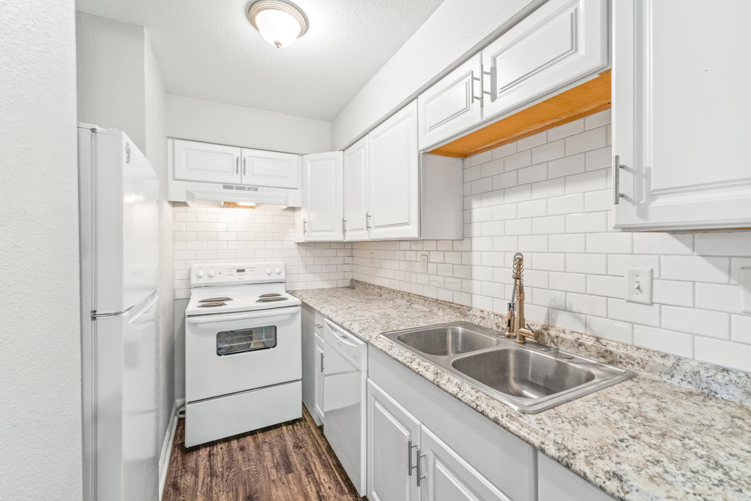 A modern kitchen featuring white cabinetry, granite countertops, a double sink, and a white electric stove. The backsplash consists of white subway tiles, and a refrigerator is positioned in the corner. Soft lighting illuminates the space, creating a bright and inviting atmosphere.