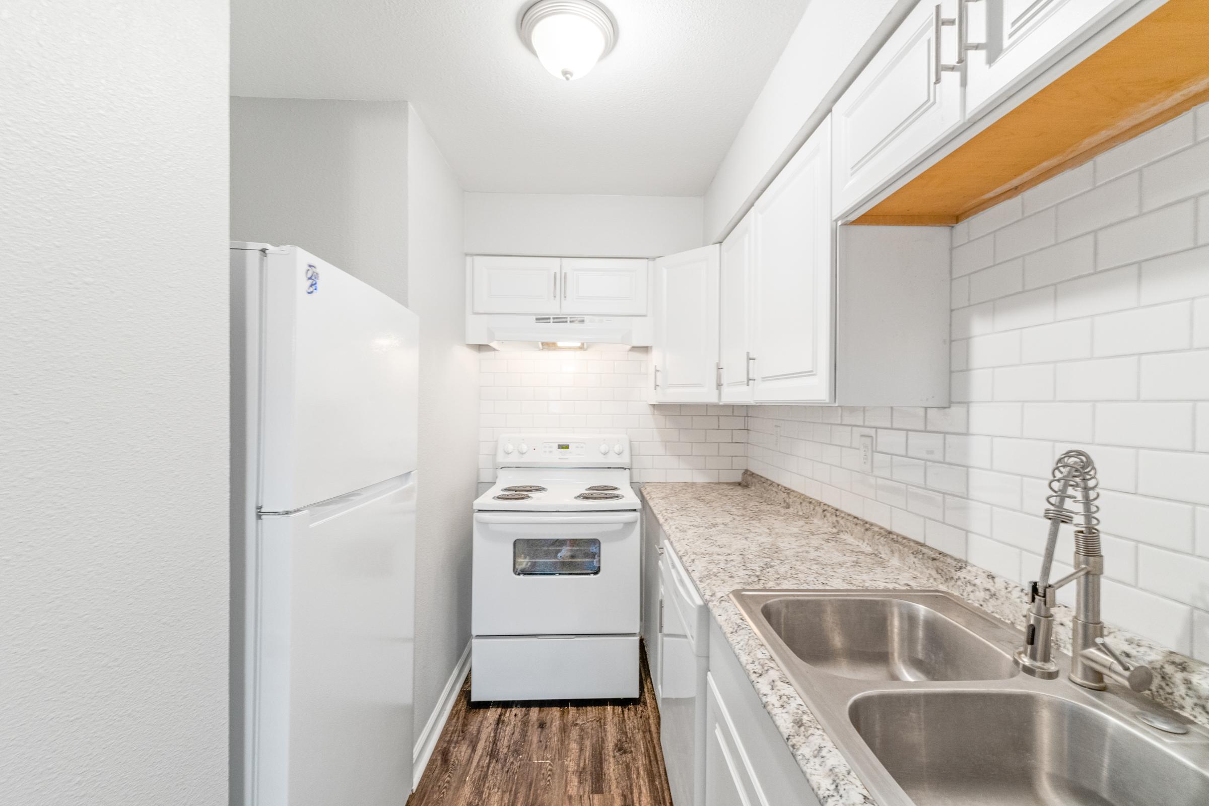 A modern kitchen featuring white cabinets, a light-colored granite countertop, a white refrigerator, and an electric stove. The backsplash is made of white tiles, and there are double stainless steel sinks with a modern faucet. The space is well-lit with overhead lighting.