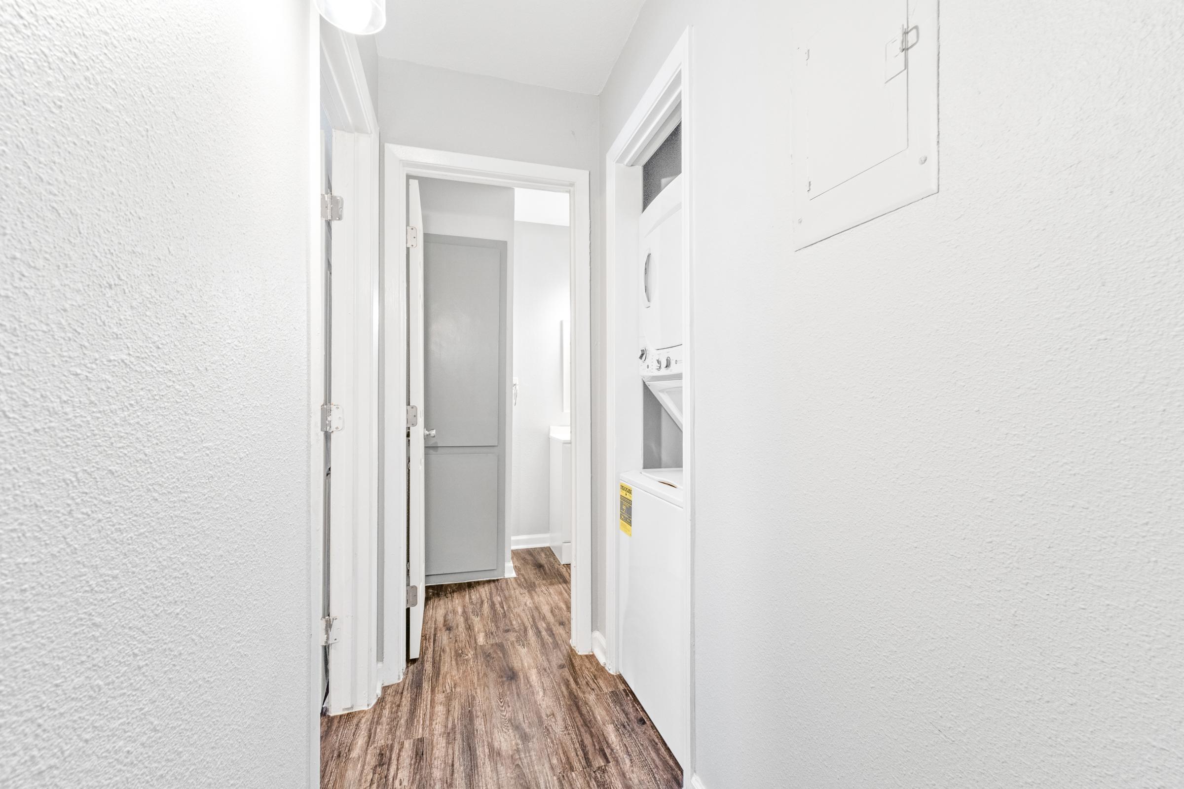 A well-lit hallway featuring light gray walls and wood-like flooring. To the left, there are two open doors leading to rooms, while a third door on the right opens to a laundry area with a stacked washer and dryer. The space appears clean and modern, with ample natural light.