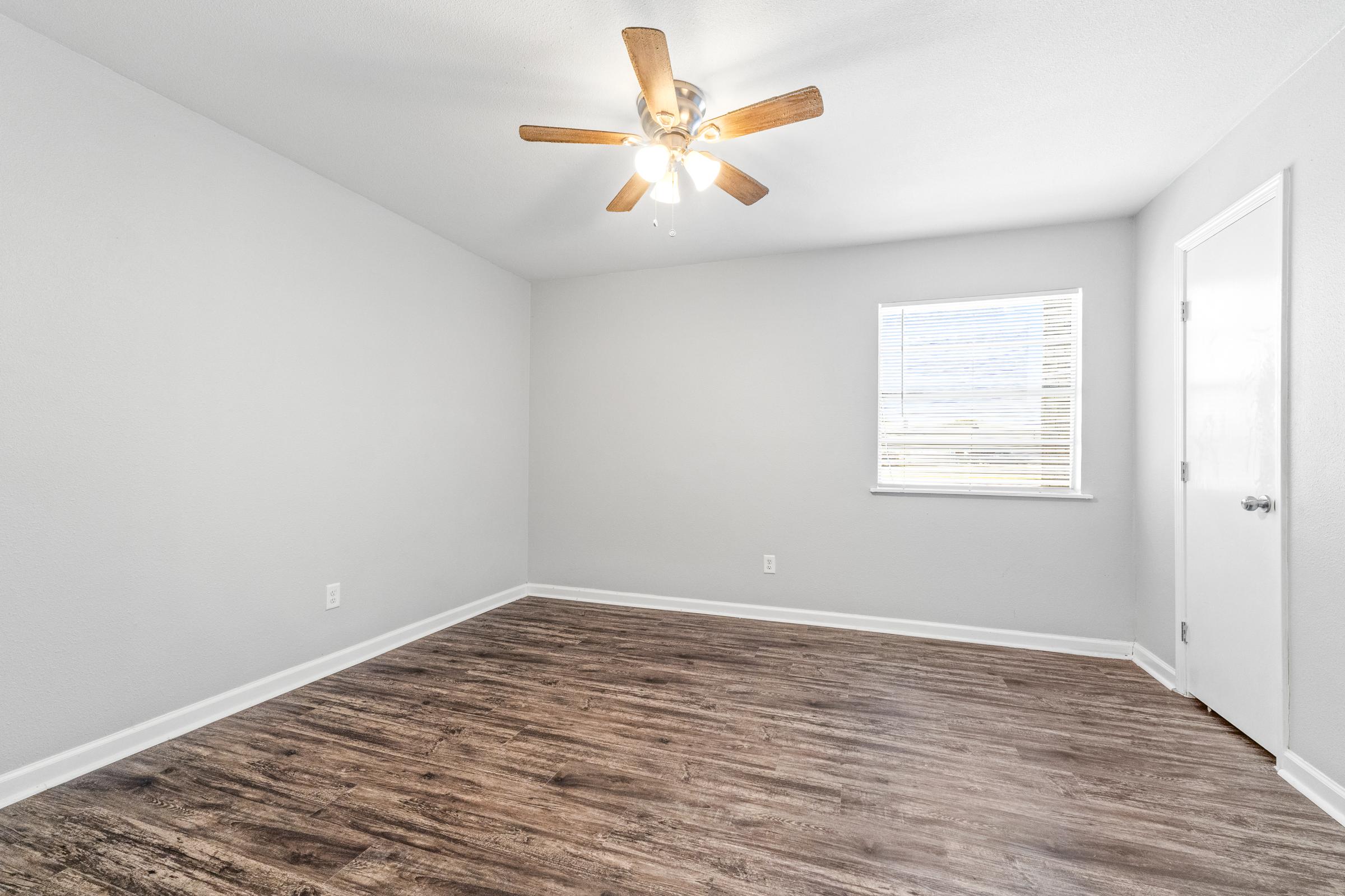 A spacious, empty room featuring light gray walls and hardwood flooring. A ceiling fan with wooden blades hangs from the ceiling. There is a single window with blinds letting in natural light, and a white door on the right side of the room. The overall atmosphere is bright and clean, ready for furnishings.