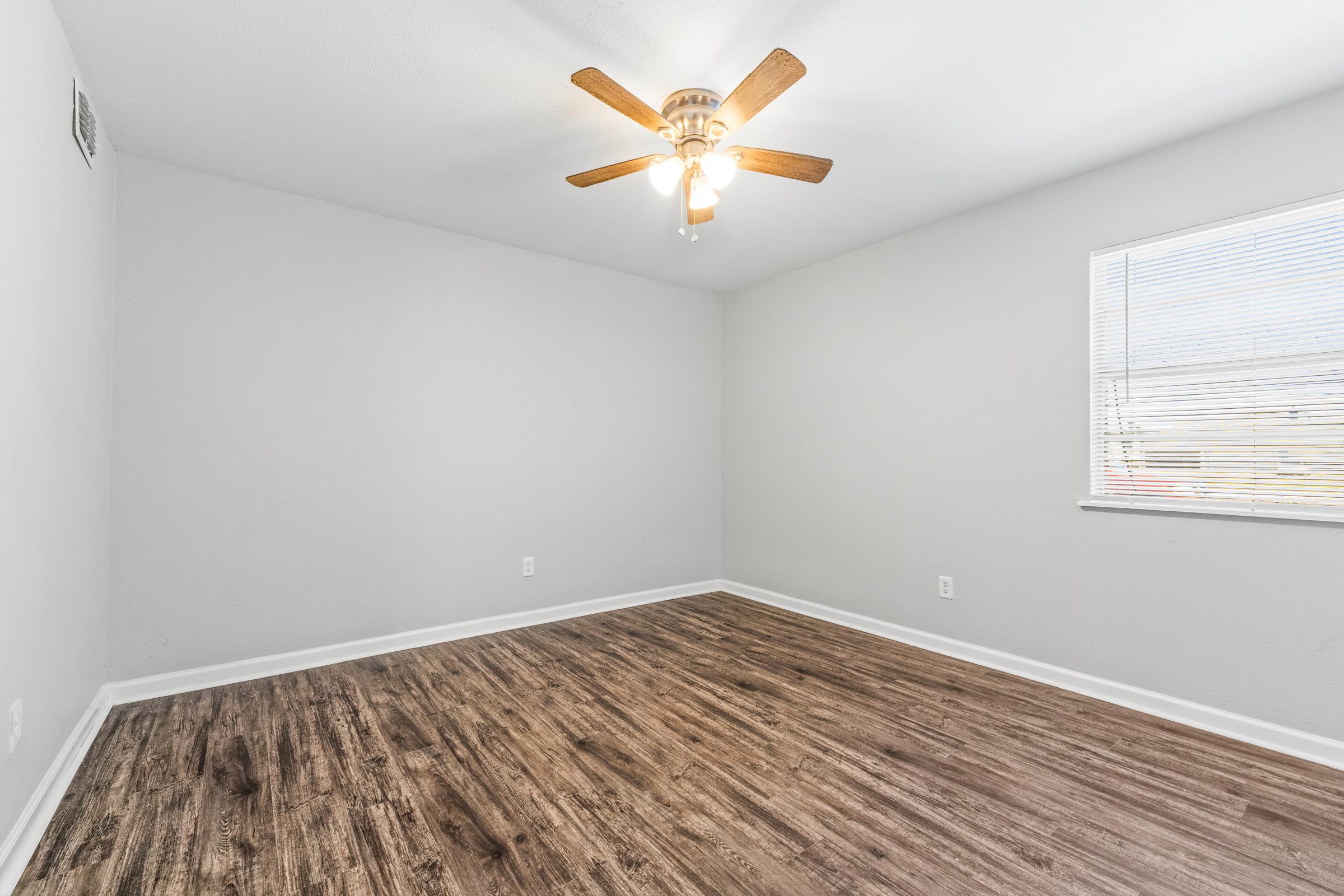 Empty room with light gray walls and a ceiling fan. The floor features brown wood laminate. A window with blinds lets in natural light, illuminating the spacious area. The room has no furniture, creating a clean and minimalist appearance.