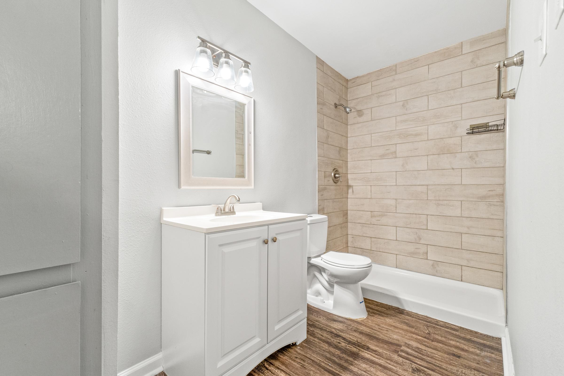 Bright, modern bathroom featuring a white vanity with a sink, a large mirror, and stylish wall sconces. The shower area has beige tiled walls, and there's a white toilet beside the vanity. The floor is wood-like laminate, creating a warm, inviting atmosphere.