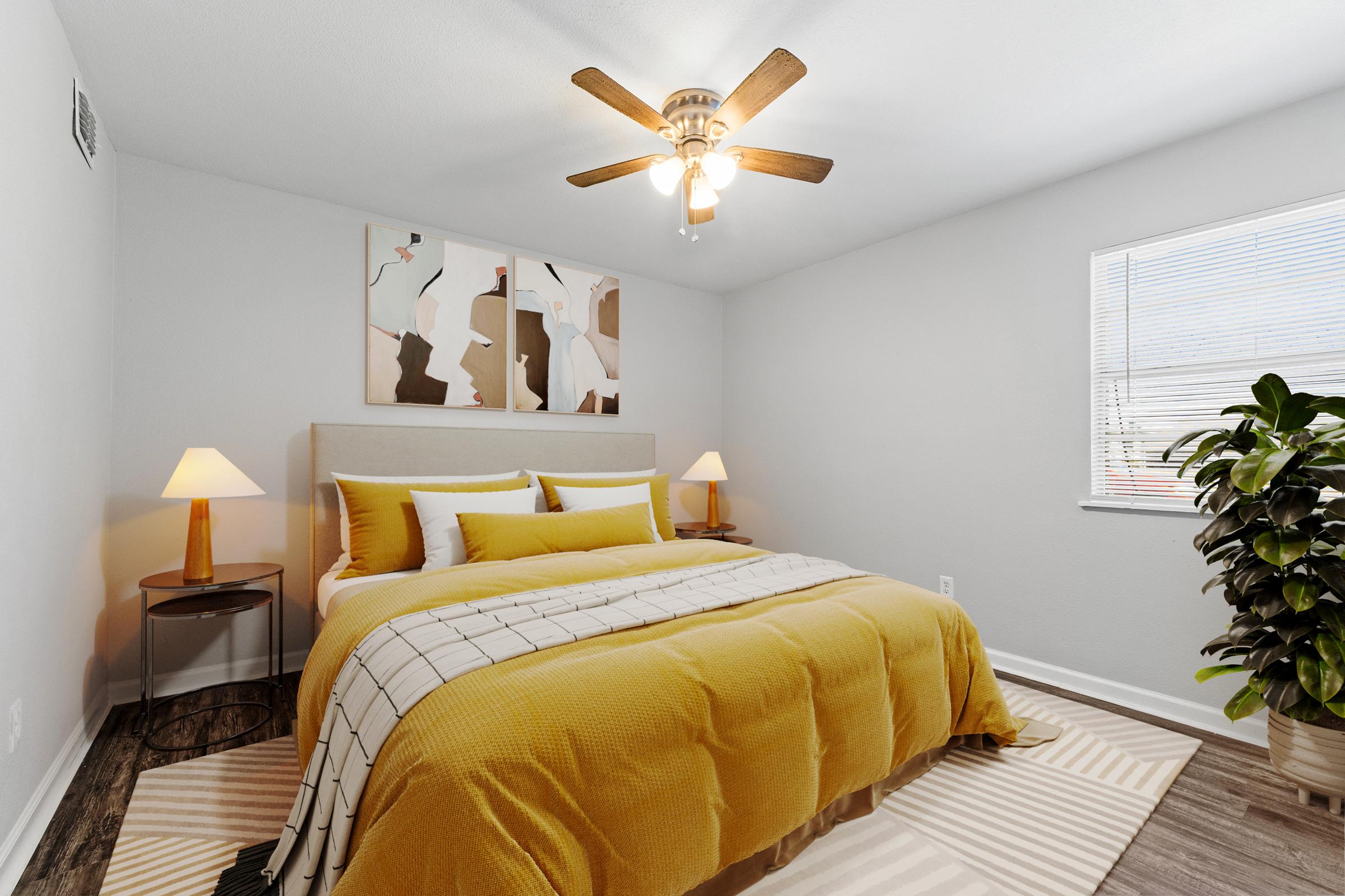 Cozy bedroom featuring a king-sized bed with yellow and white bedding, a modern ceiling fan, and warm lighting from two bedside lamps. Decorative artwork hangs above the bed, and a potted plant adds a touch of greenery. The walls are painted light gray, and a striped area rug complements the decor.