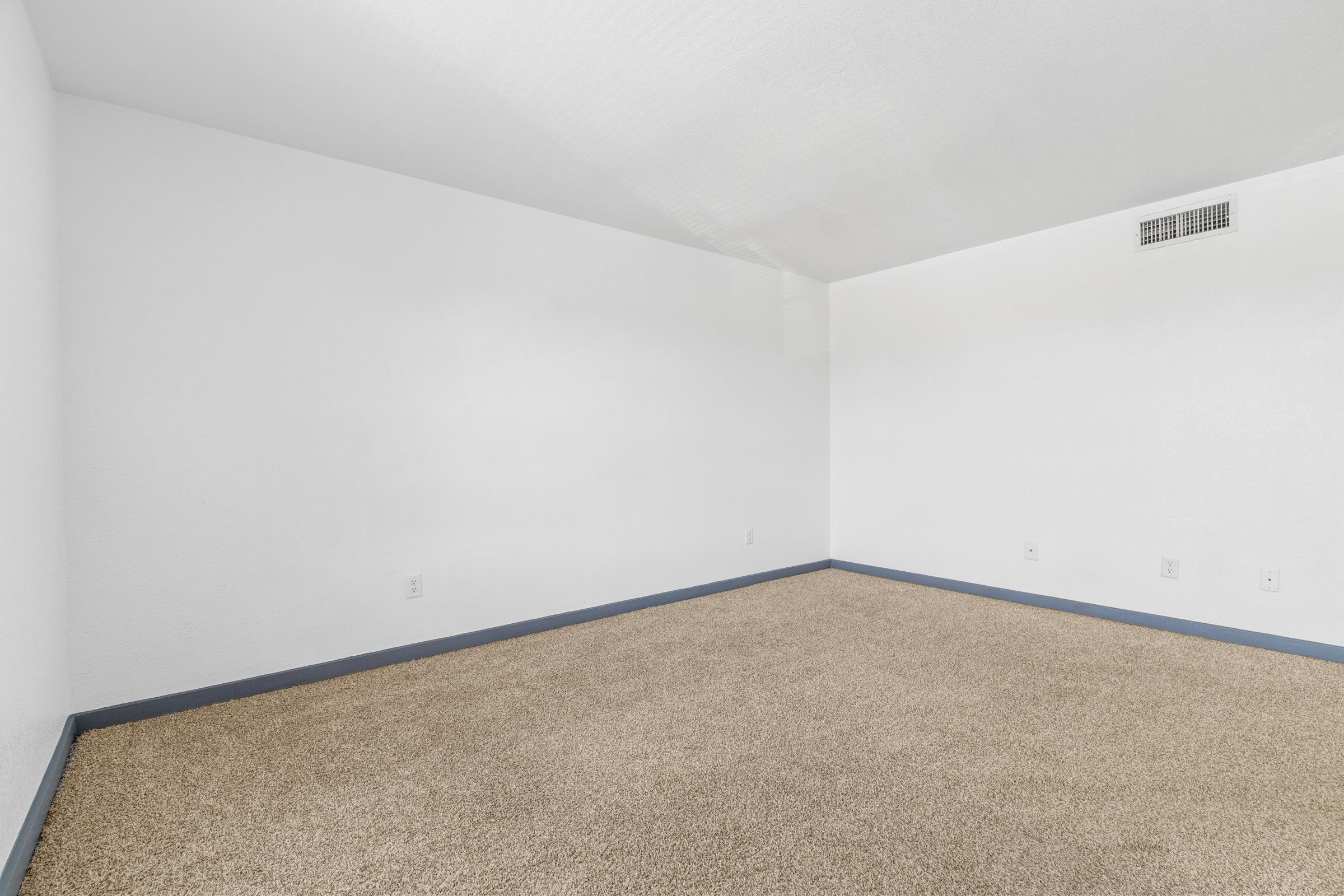 A spacious, empty room featuring light gray walls and a soft beige carpet. The room has no furniture and is well-lit, with one wall having a vent. The corners are clean and the overall space appears inviting for various uses or interior decoration.