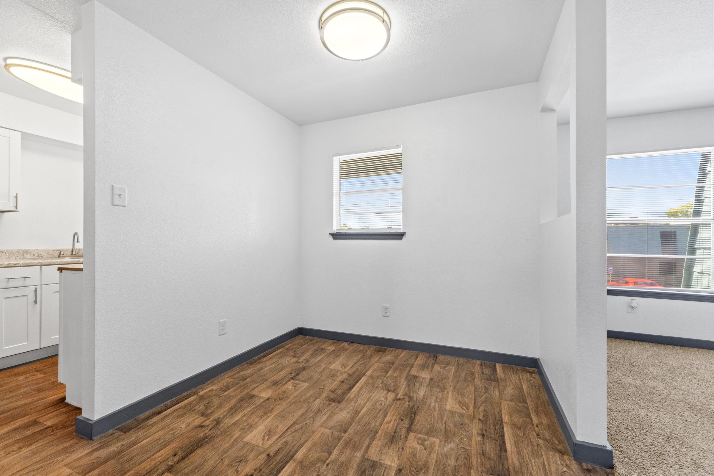 A clean, modern interior of a room featuring a hardwood floor, white walls, and a ceiling light. There's a window on the left with blinds, and a glimpse of another room visible through an open doorway on the right. The space appears bright and well-lit.