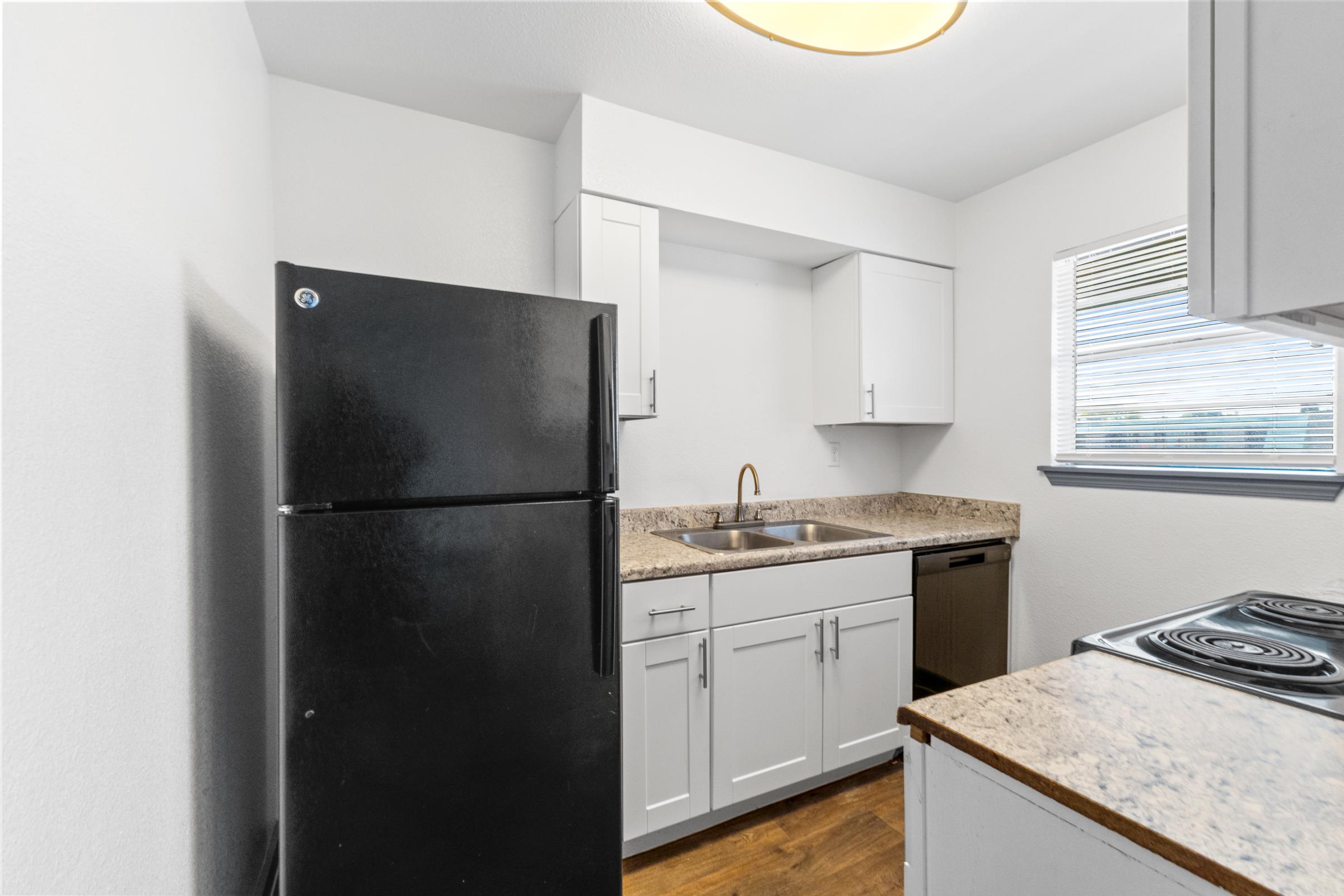 A modern kitchen featuring a black refrigerator, white cabinetry, a stainless steel sink, and a countertop made of speckled granite. There’s a stove with an electric range and a window that lets in natural light, illuminating the space with a minimalist design.