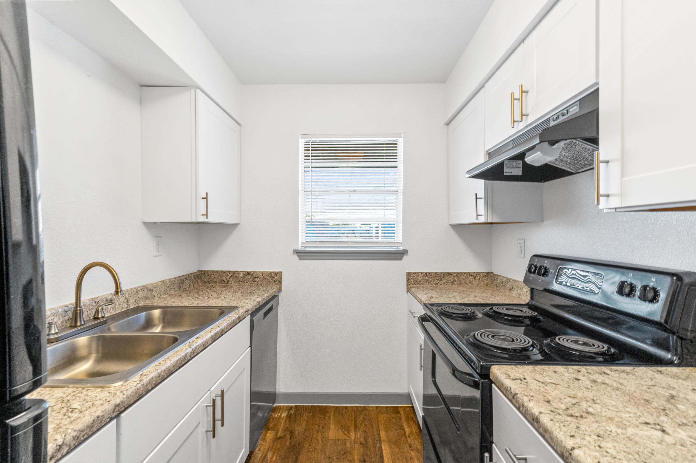 Modern kitchen featuring white cabinetry, a double sink with a gold faucet, and a black stove and refrigerator. Countertops are a textured light stone. A window provides natural light, illuminating the space. The floor is a warm wood tone, adding a cozy touch to the design.